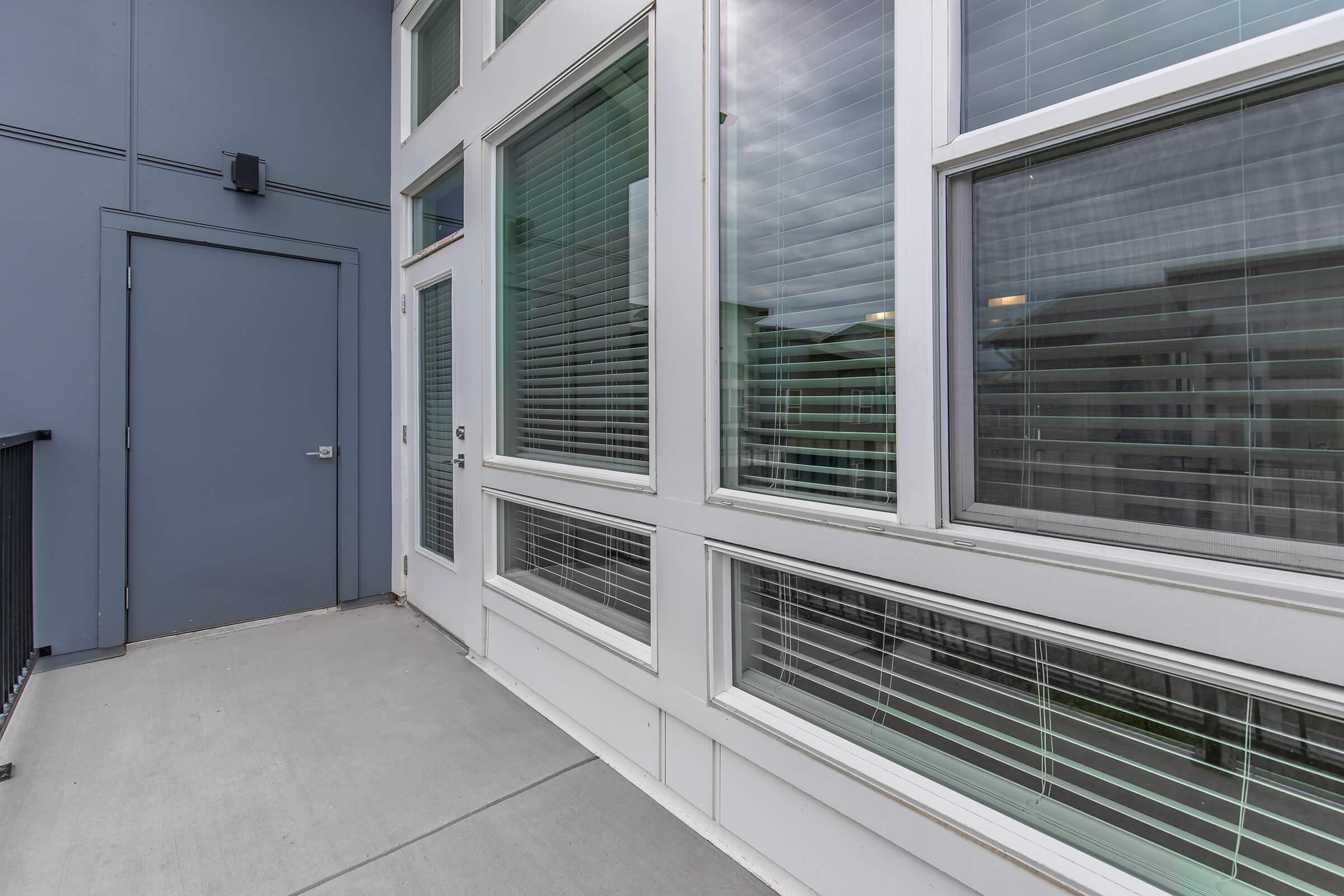 A modern building exterior featuring large windows with horizontal blinds and a gray door. The surface is a concrete walkway leading up to the door, with a gray wall on the left side and a view of the windows on the right. The overall design is sleek and contemporary.