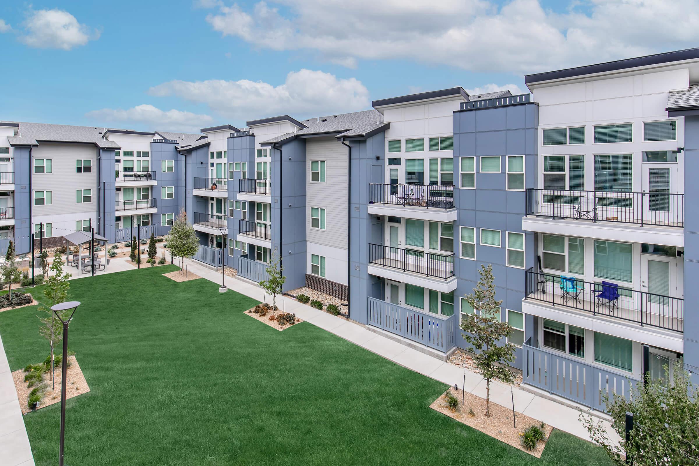A modern apartment complex with three-story buildings featuring a mix of gray and white exteriors. Each building has balconies and large windows. The foreground includes a well-maintained green lawn and landscaped areas with small trees and walkways, under a partly cloudy blue sky.