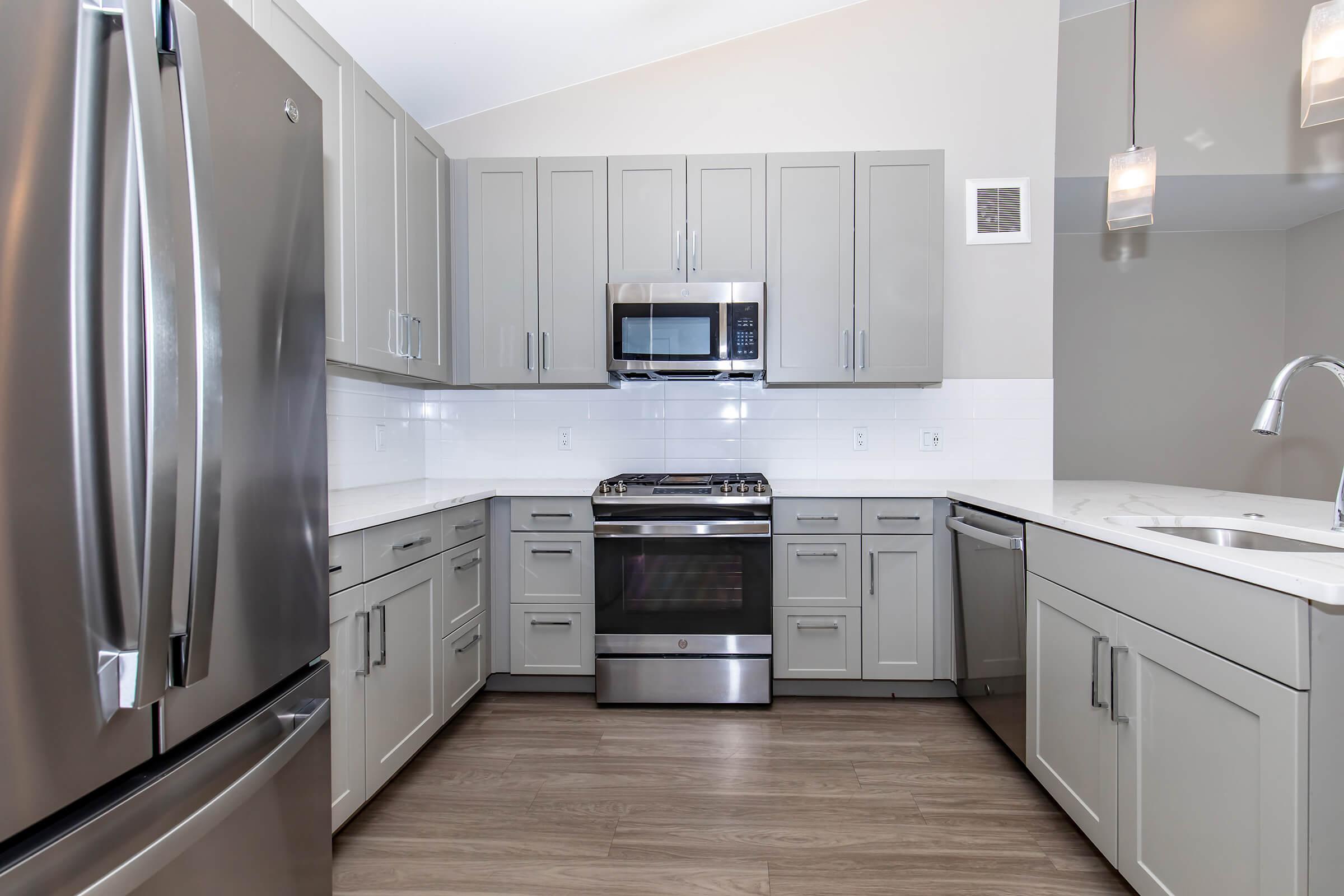 A modern kitchen featuring gray cabinets, stainless steel appliances including a refrigerator, oven, and microwave. The countertop is white, and there is a silver kitchen faucet. The floor is light-colored wood. Soft pendant lights hang from the ceiling, illuminating the space.