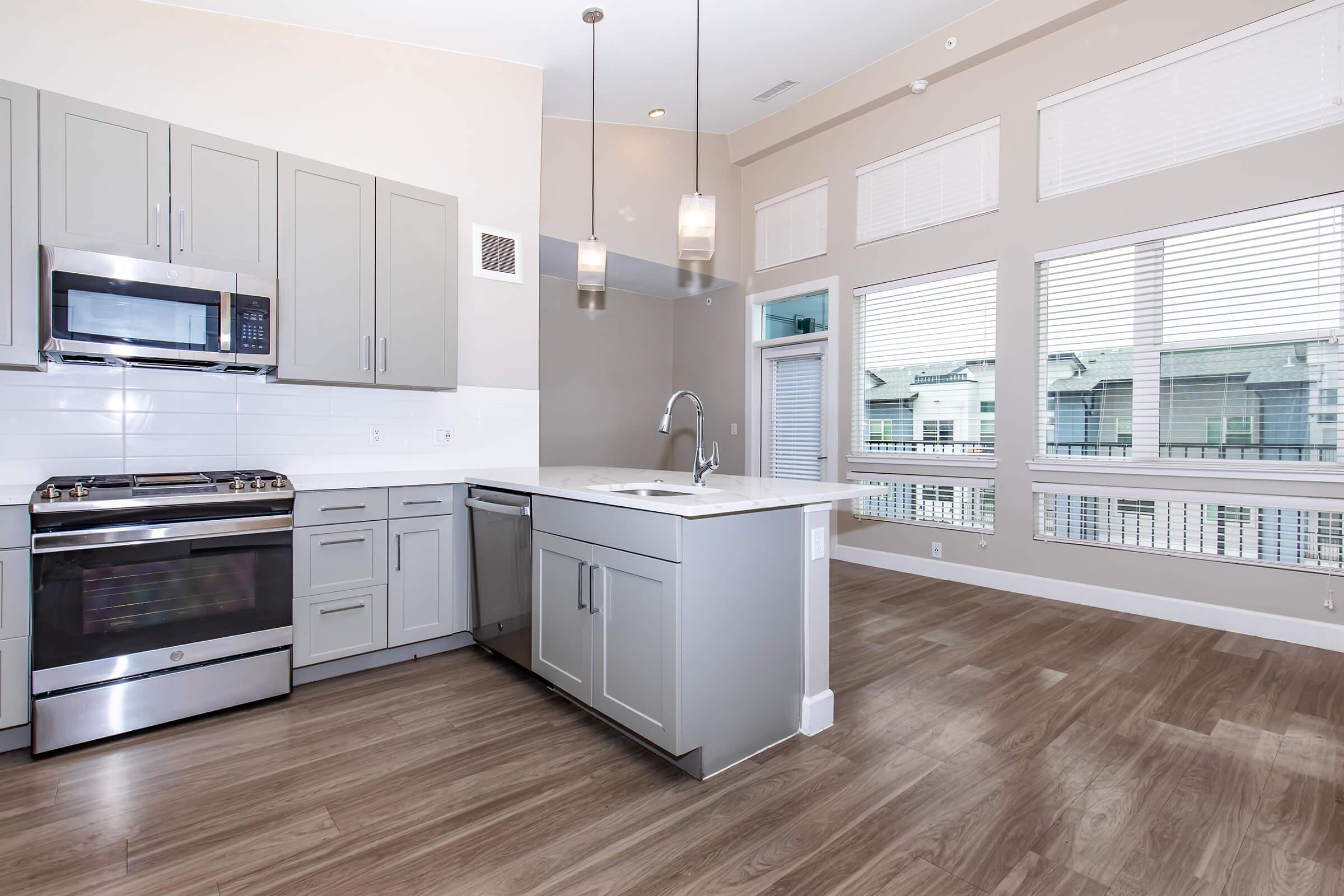 Modern kitchen with gray cabinetry, stainless steel appliances, and a white countertop. Features large windows allowing natural light, hardwood-style flooring, and pendant lighting. The layout is open and airy, creating a spacious feel in the living area.