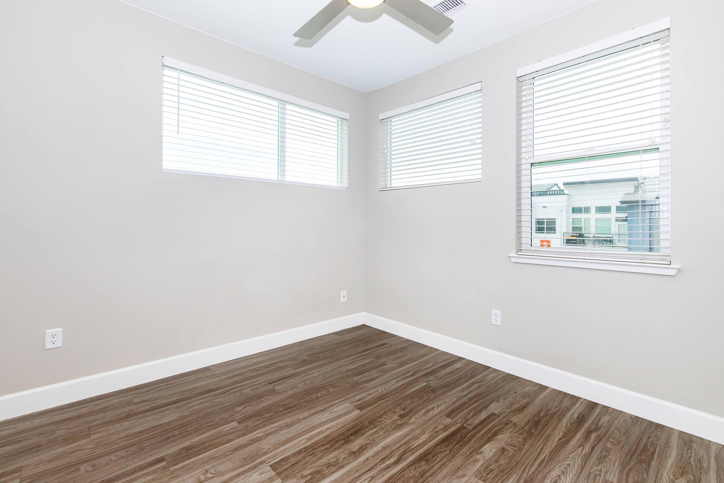 Empty room featuring light-colored walls, a ceiling fan, and multiple windows with blinds. The flooring is a wood-like laminate. Natural light fills the space, highlighting the simplicity and modern design of the room.
