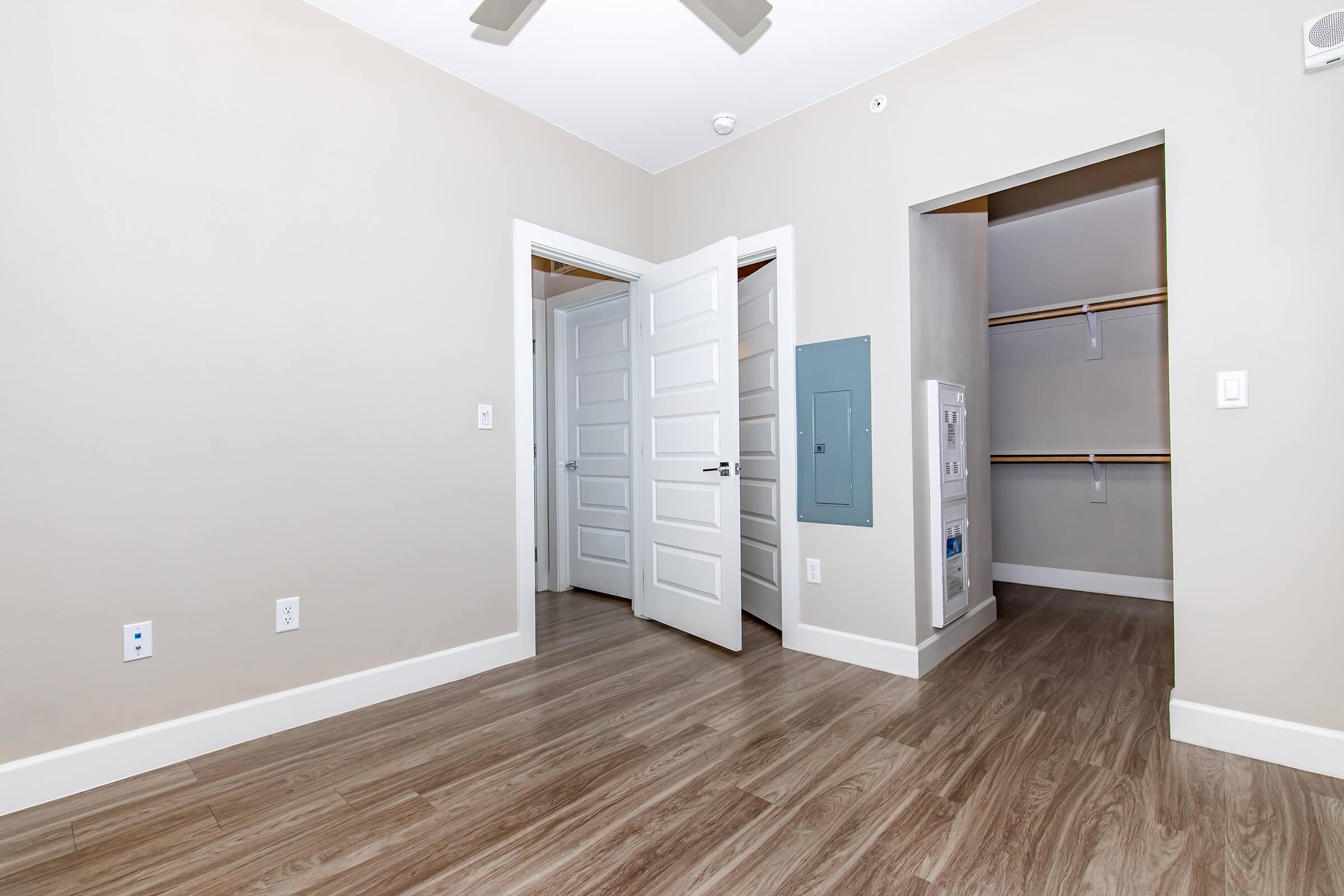 A light-colored room with wooden flooring features two open white doors leading to a closet and another room. A ventilation fan is visible on the ceiling, and there is a utility panel on the wall beside the closet. The walls are painted in a neutral shade, contributing to a bright and spacious atmosphere.