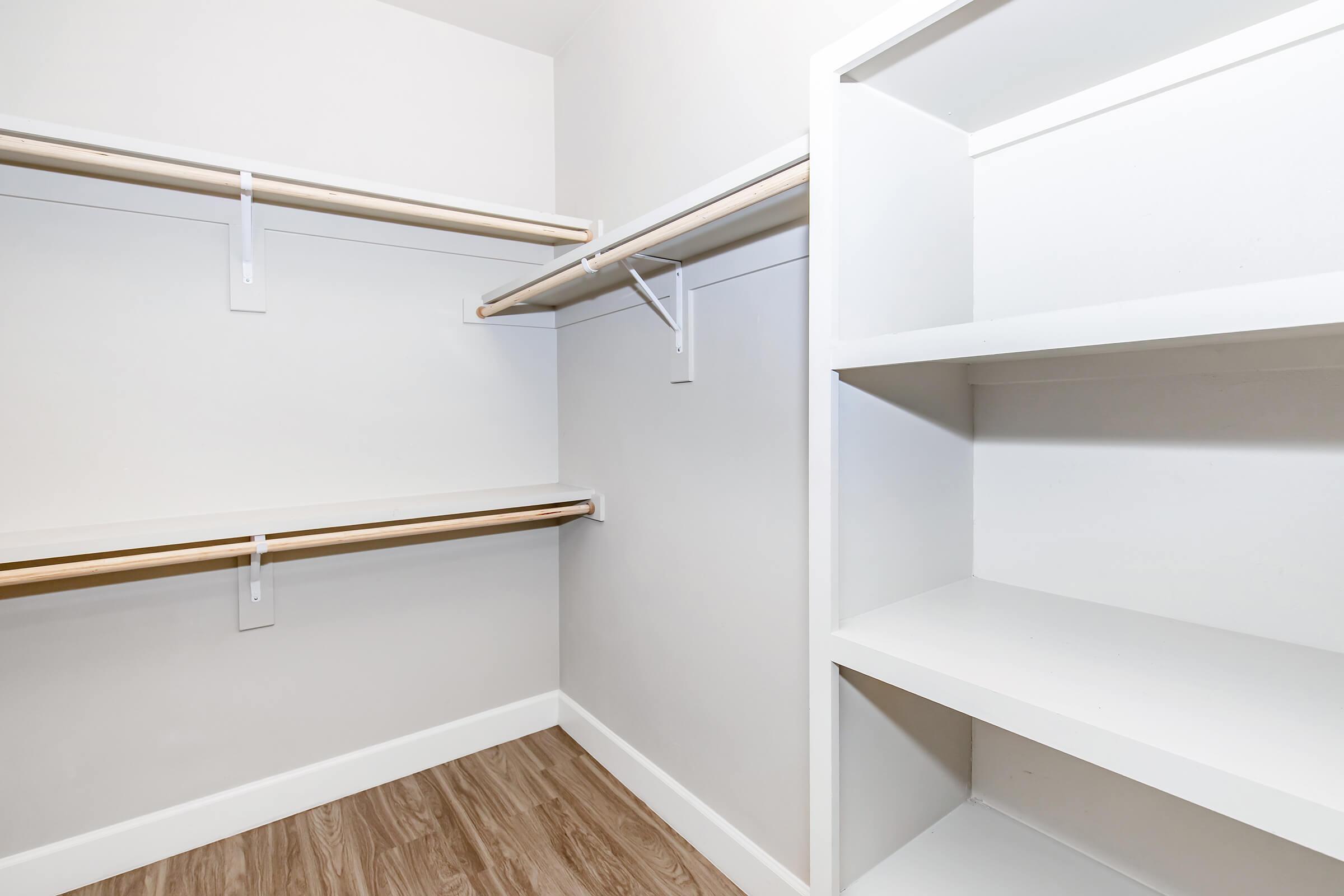 A clean and empty closet space featuring two hanging rods on the left side and a shelf on the right side, all set against a light-colored wall. The flooring is a light wood finish, contributing to a bright and organized appearance.