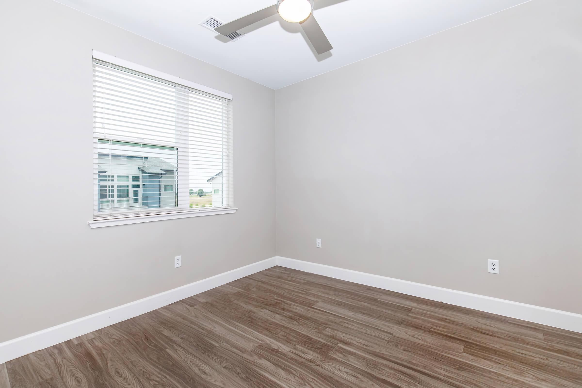An empty room with light-colored walls and vinyl flooring. It features a ceiling fan and a window with blinds, providing natural light. The room appears spacious and minimalistic, suitable for various uses like an office or bedroom.