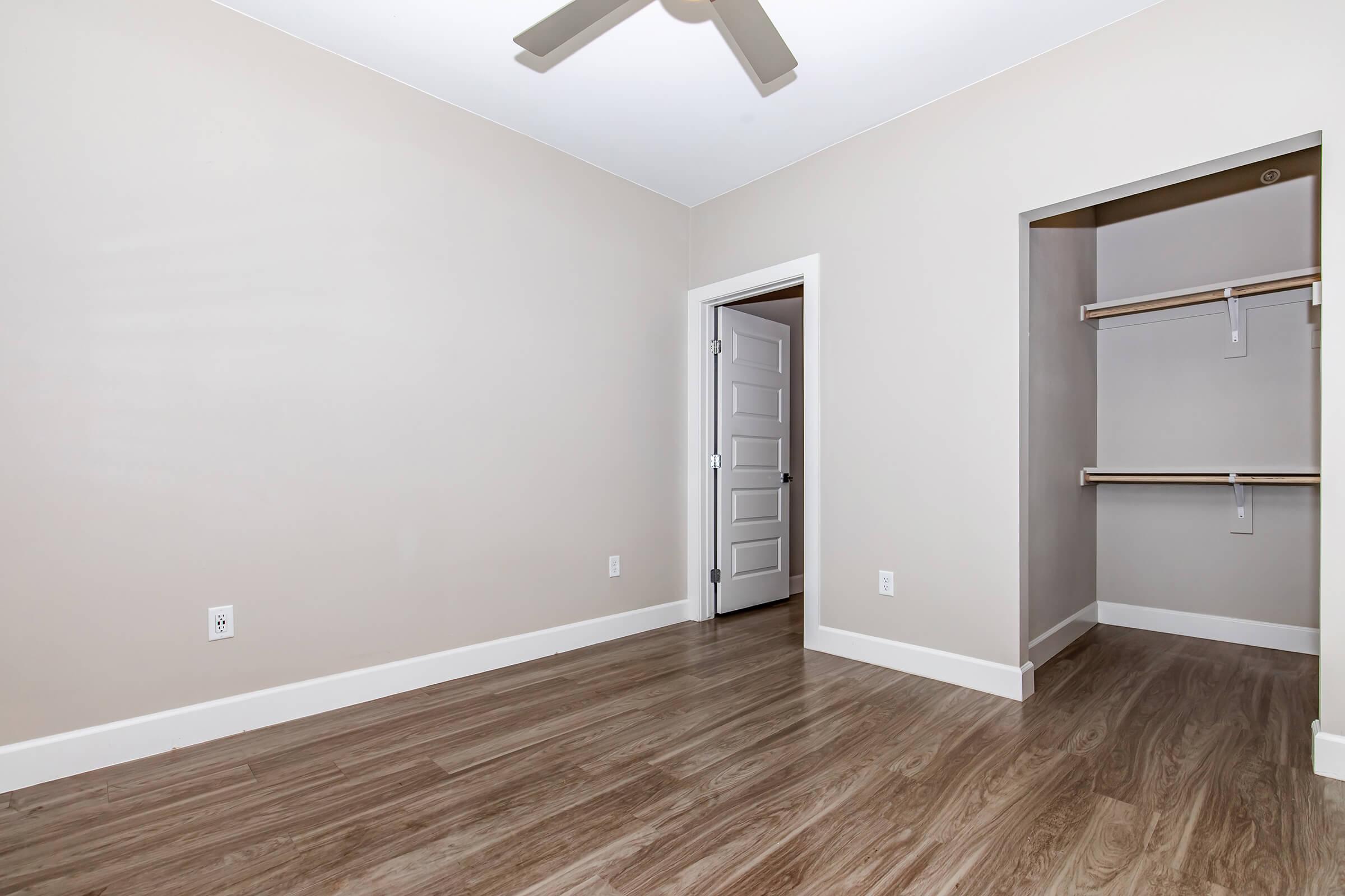 Empty room with light-colored walls and a ceiling fan. There is a white door on the left leading to another space and an adjacent closet with wooden shelves. The flooring is a light, wood-like material, contributing to a clean, minimalist aesthetic.
