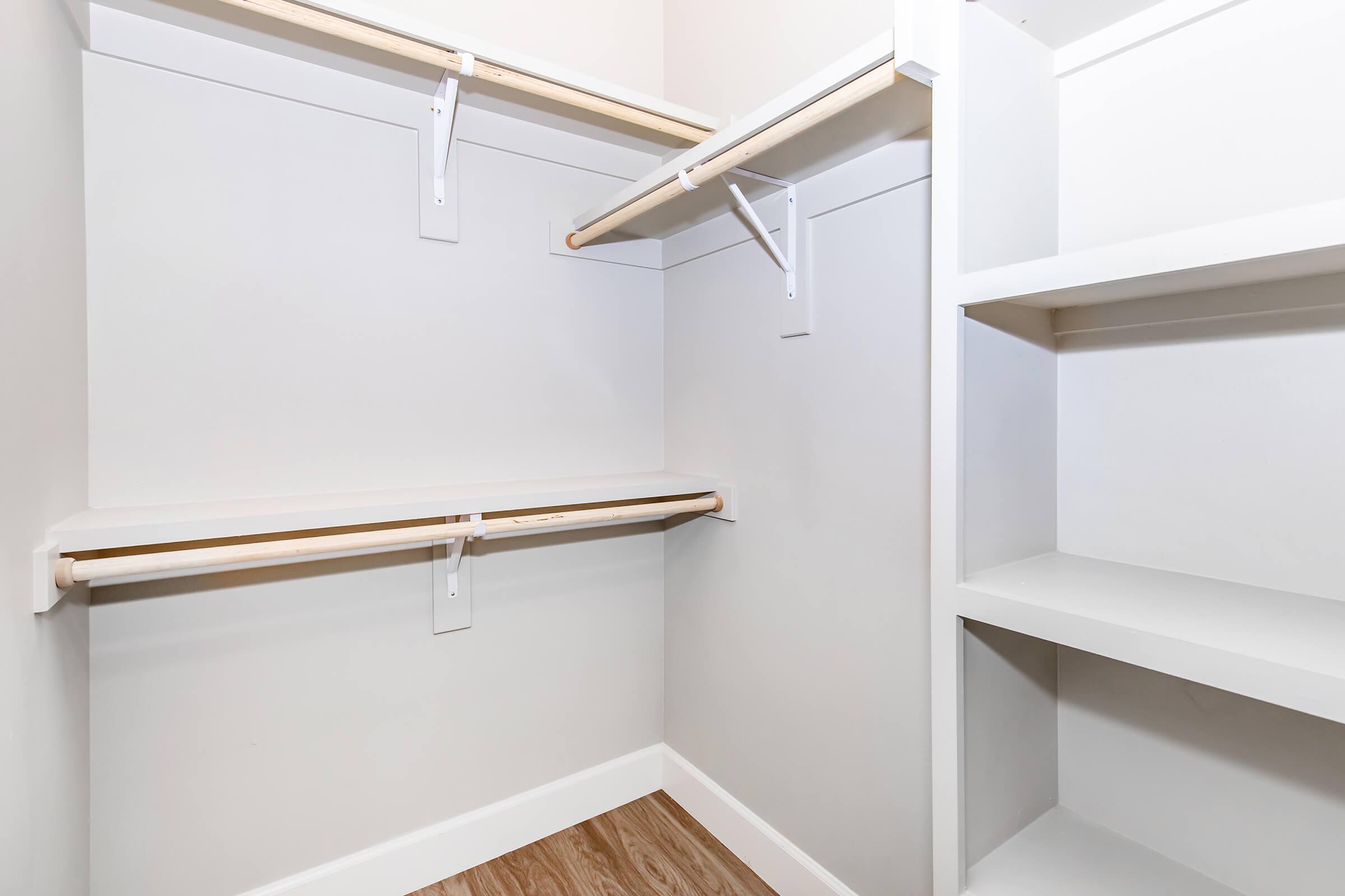 A spacious interior of a walk-in closet featuring two wooden shelves supported by brackets, with a clean, minimal design. The walls are light-colored, and there is a section with open shelving, which is also empty, providing ample storage possibilities. The flooring is wooden.