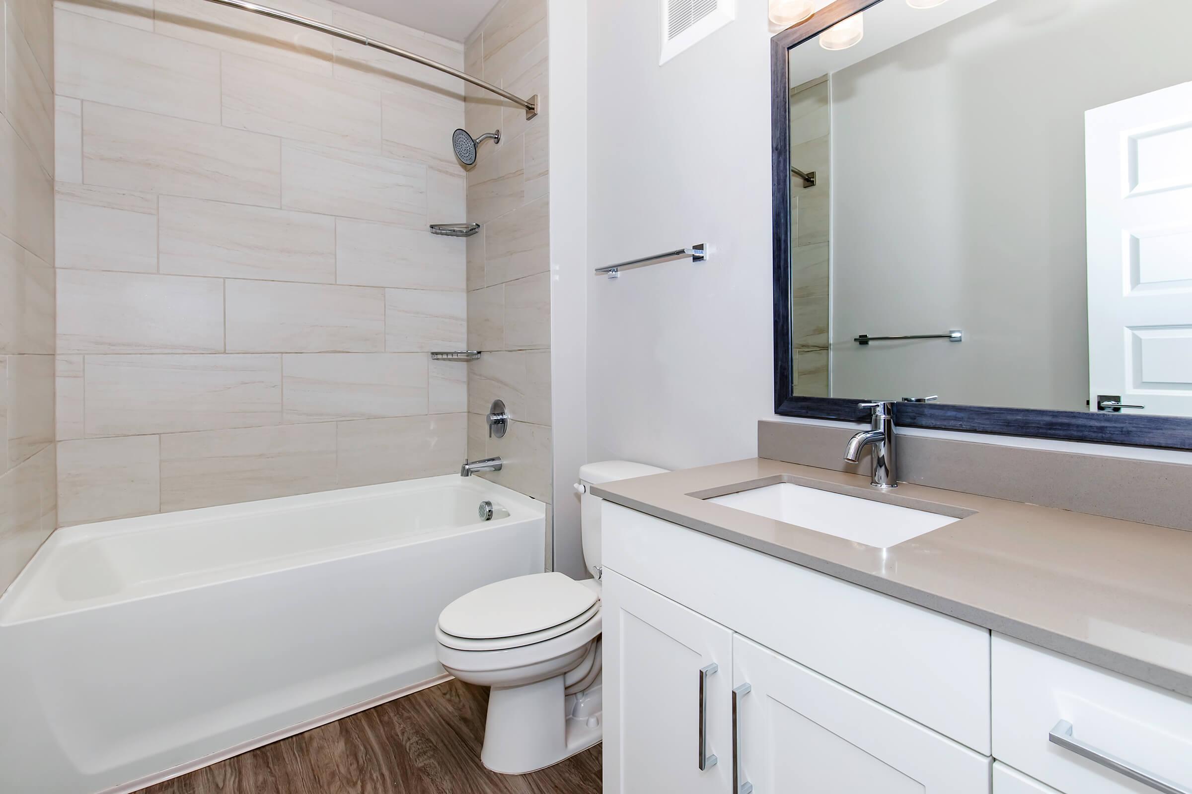 A modern bathroom featuring a bathtub with a showerhead, a white toilet, and a double vanity with a rectangular sink. The walls are tiled with light-colored tiles, and there is a large mirror above the vanity. The flooring is dark wood, creating a contrast with the light fixtures and walls.