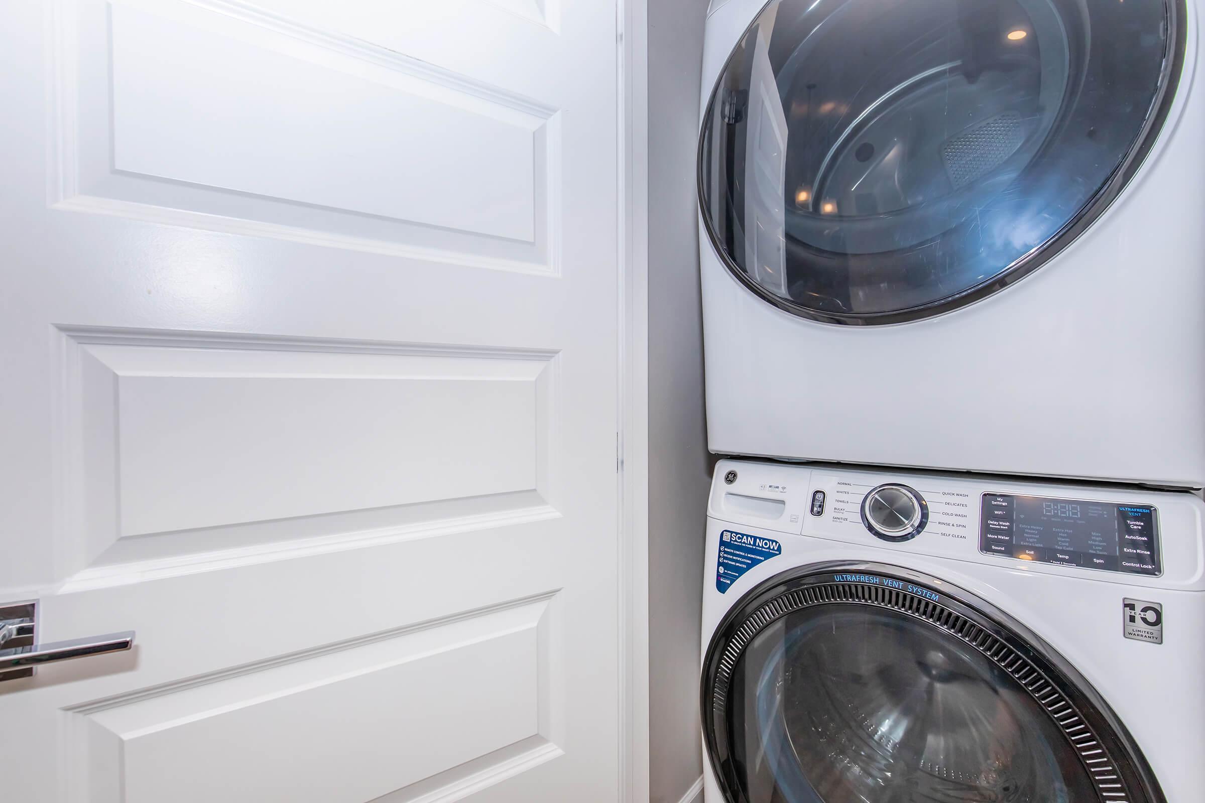 A modern stacked washer and dryer unit situated next to a closed white door. The appliances are sleek and feature a digital display panel, showcasing a clean and organized laundry area. Soft lighting highlights the shiny surfaces of the machines.