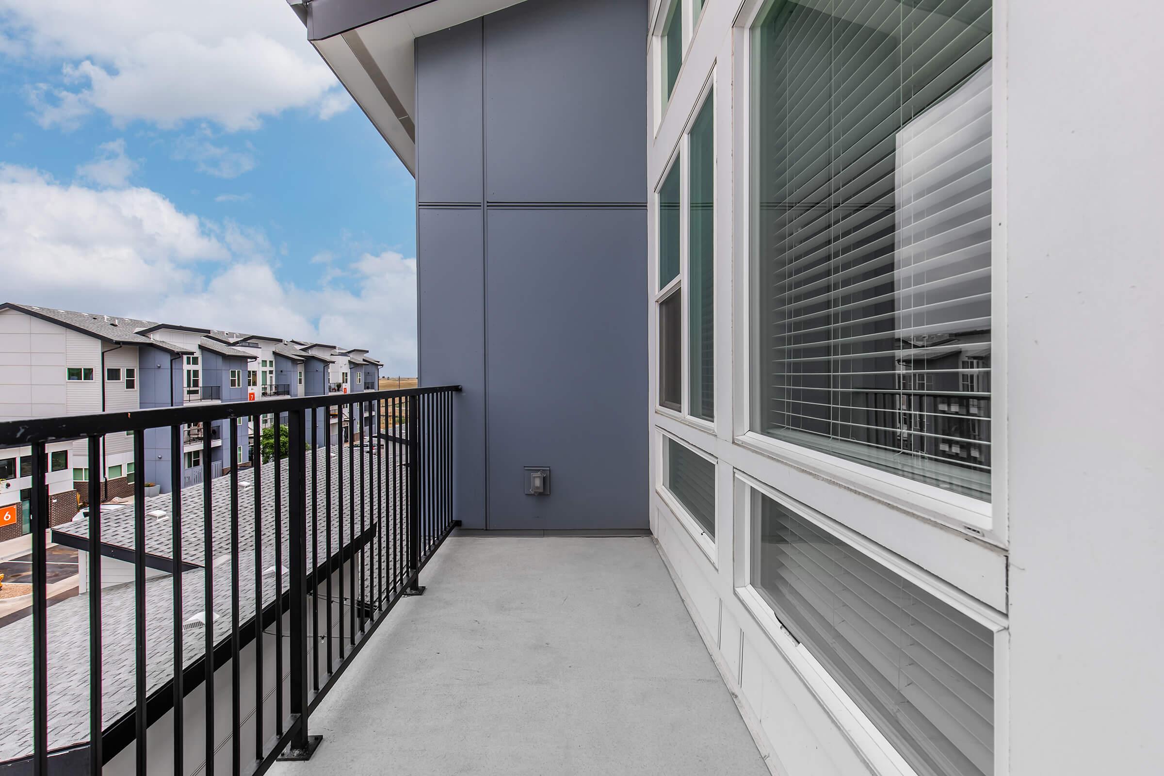 A spacious balcony with a black railing, featuring a smooth concrete floor. The background shows the exterior of neighboring buildings and a clear blue sky with scattered clouds. Large windows provide a view of the outside.