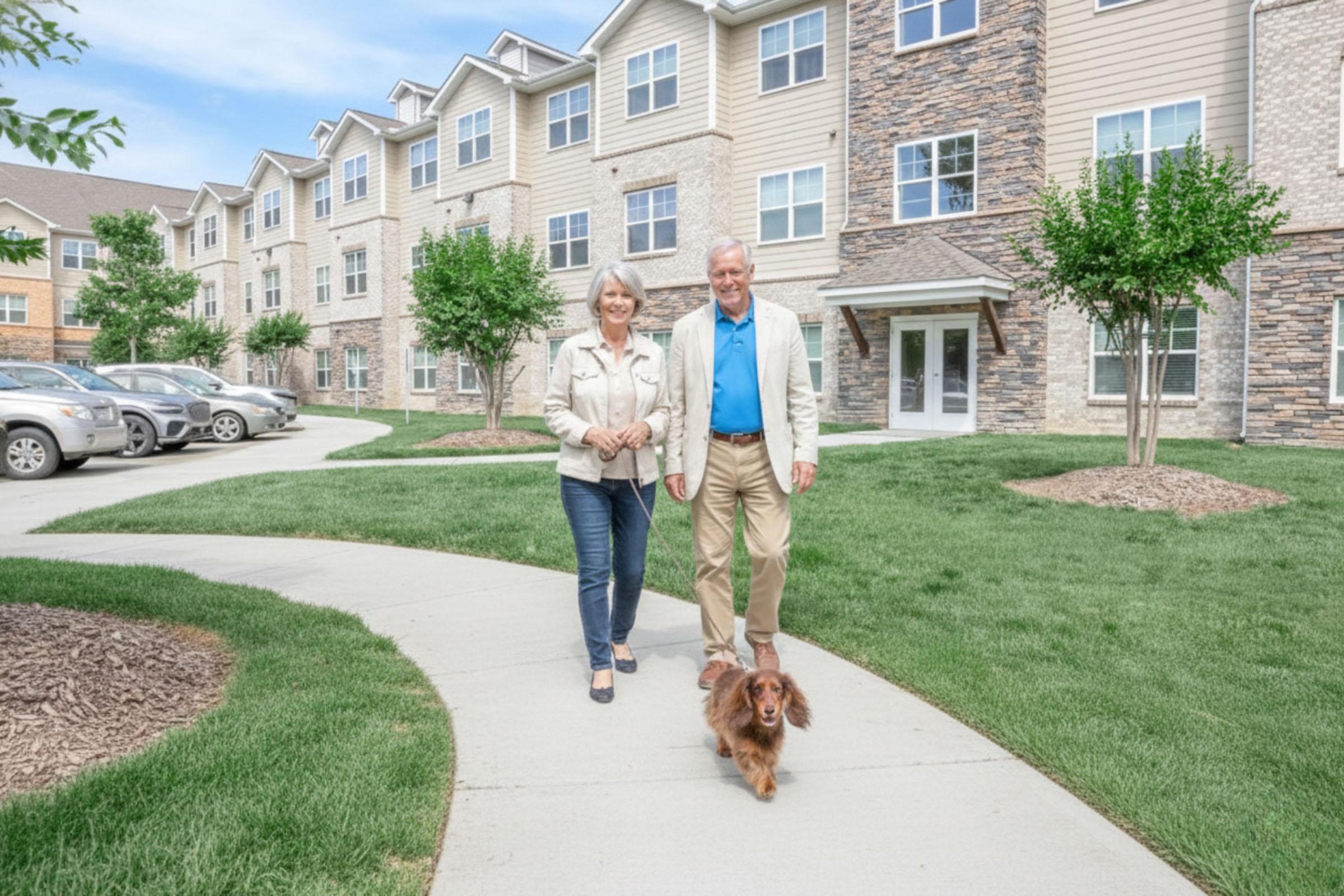 A senior couple walks together on a path in front of a multi-story apartment building, accompanied by a small dog. The setting features well-manicured grass and trees, with parked cars nearby. The sky is clear, suggesting a pleasant day.