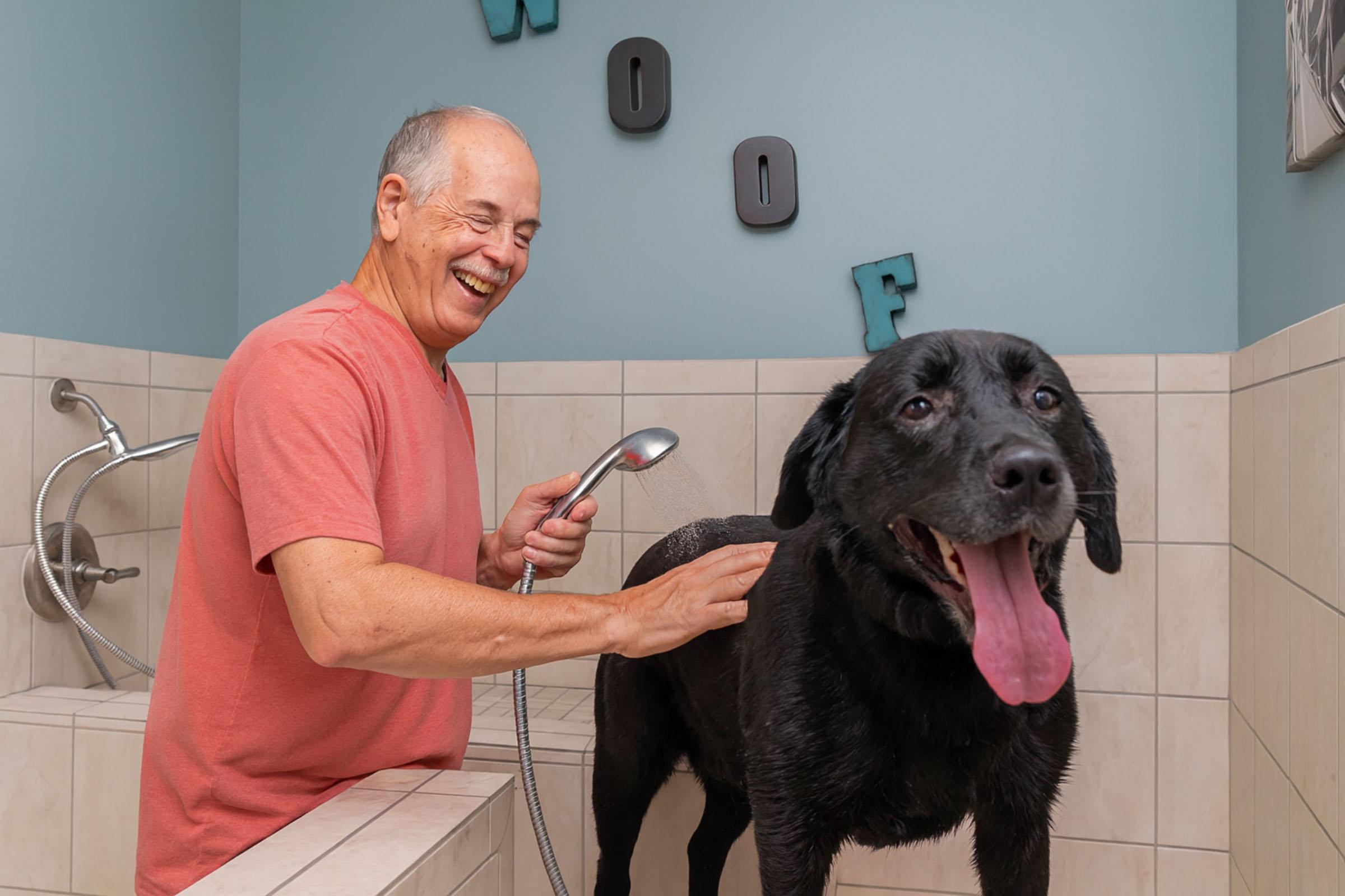 A smiling older man bathes a large black dog in a tiled shower area. The dog, looking happy with its tongue out, stands on a platform while the man gently holds a handheld showerhead, spraying water. The wall in the background has the word "WOOF" displayed in playful letters.