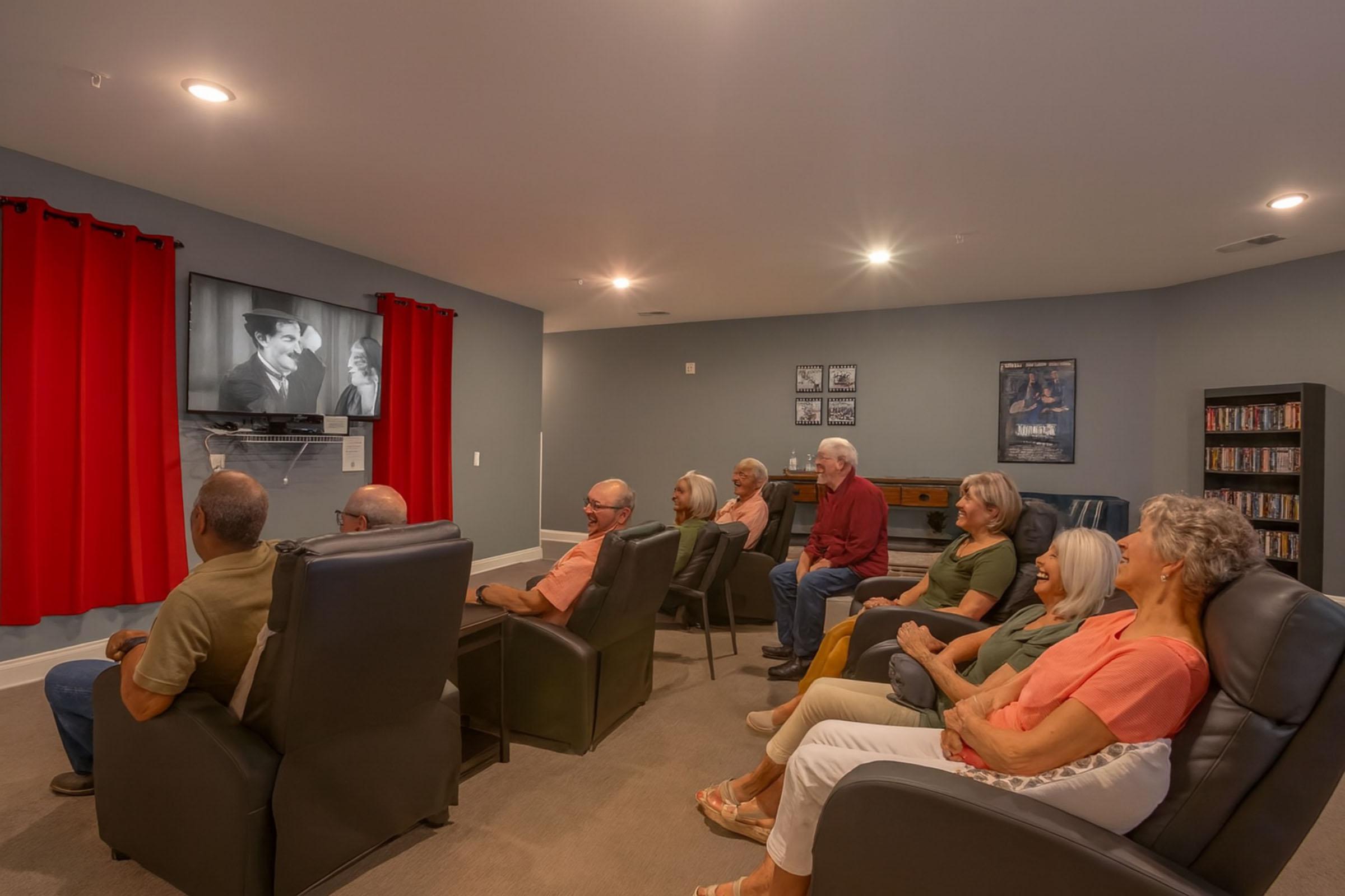 A cozy home theater room with eight adults seated in comfortable chairs, watching a black-and-white film on a wall-mounted screen. The room has red curtains, a gray color scheme, and shelves with DVDs, creating a relaxed atmosphere for movie watching.