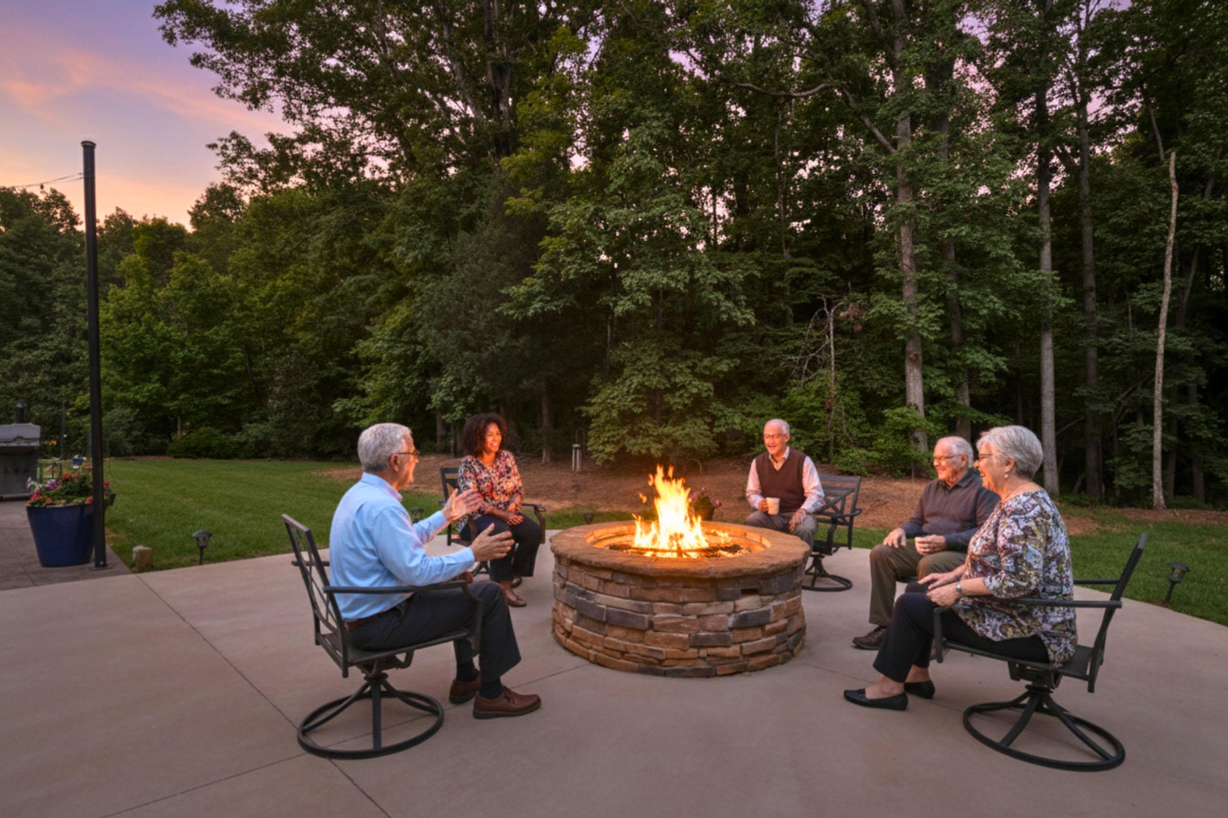 A group of five people sits around a stone fire pit during sunset, surrounded by trees. They are engaged in conversation, with warm light from the fire illuminating the scene. The setting features a patio area with chairs, creating a cozy outdoor gathering atmosphere.