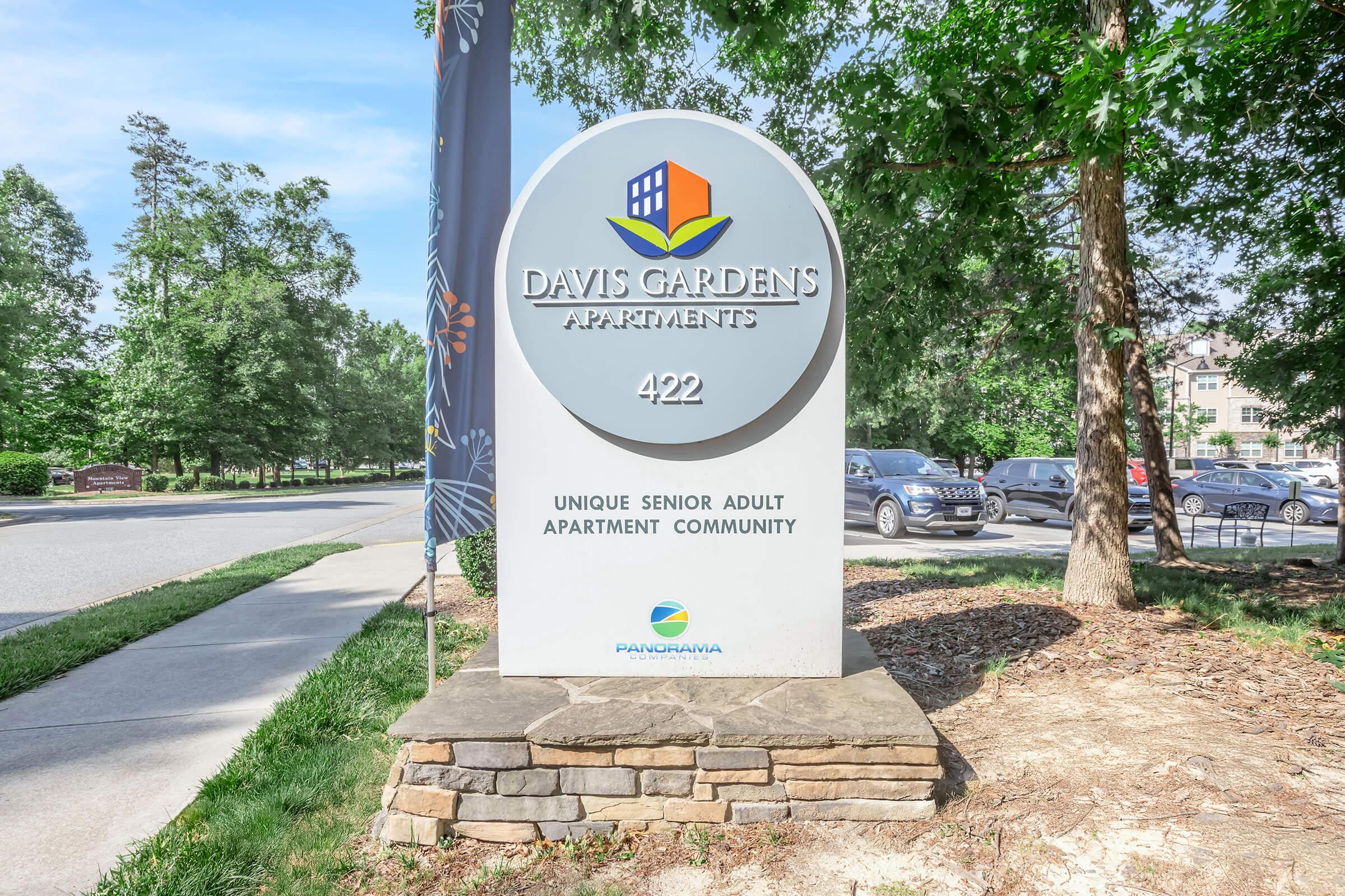 Sign for Davis Gardens Apartments, stating "Davis Gardens Apartments," with the text "422" and "Unique Senior Adult Apartment Community." The sign is situated in a landscaped area with trees and a road nearby.