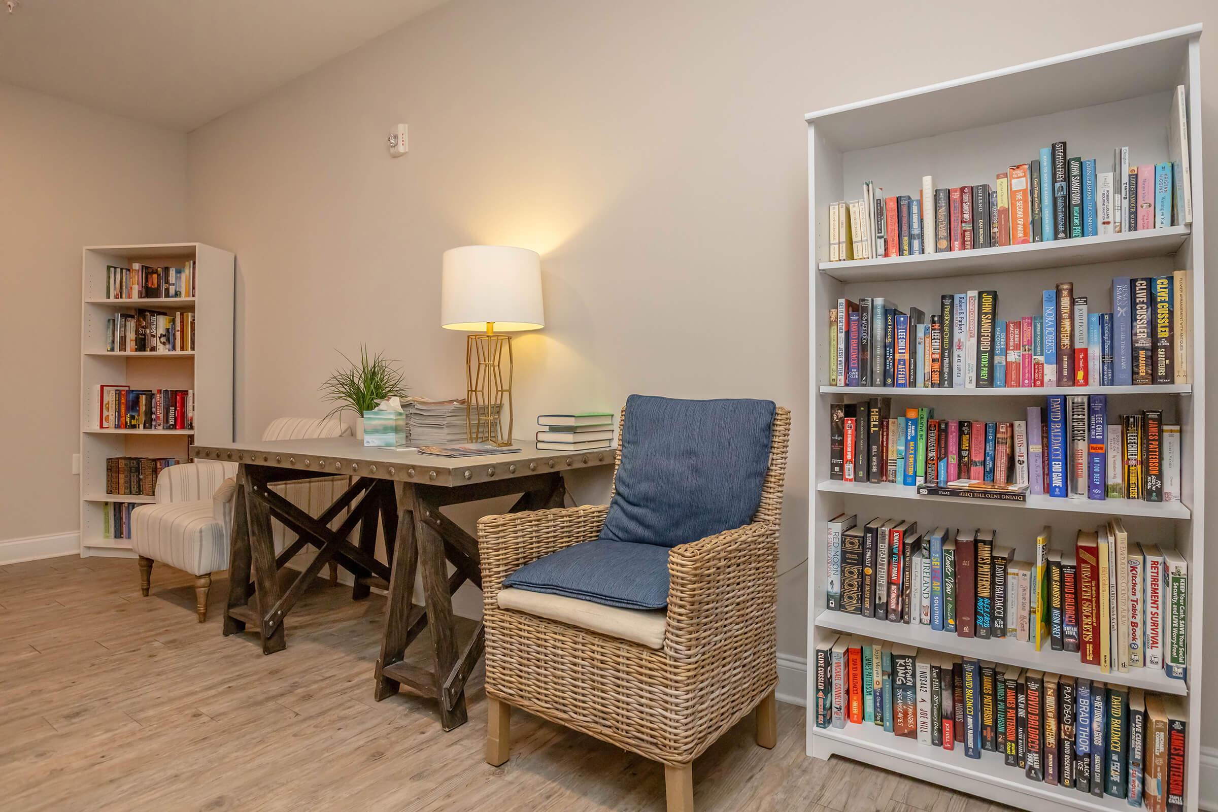 A cozy reading nook featuring a wooden table with two upholstered chairs, surrounded by bookshelves filled with various books. A decorative lamp provides warm lighting, and a small plant adds a touch of greenery to the space. The flooring is light wood, enhancing the inviting atmosphere.