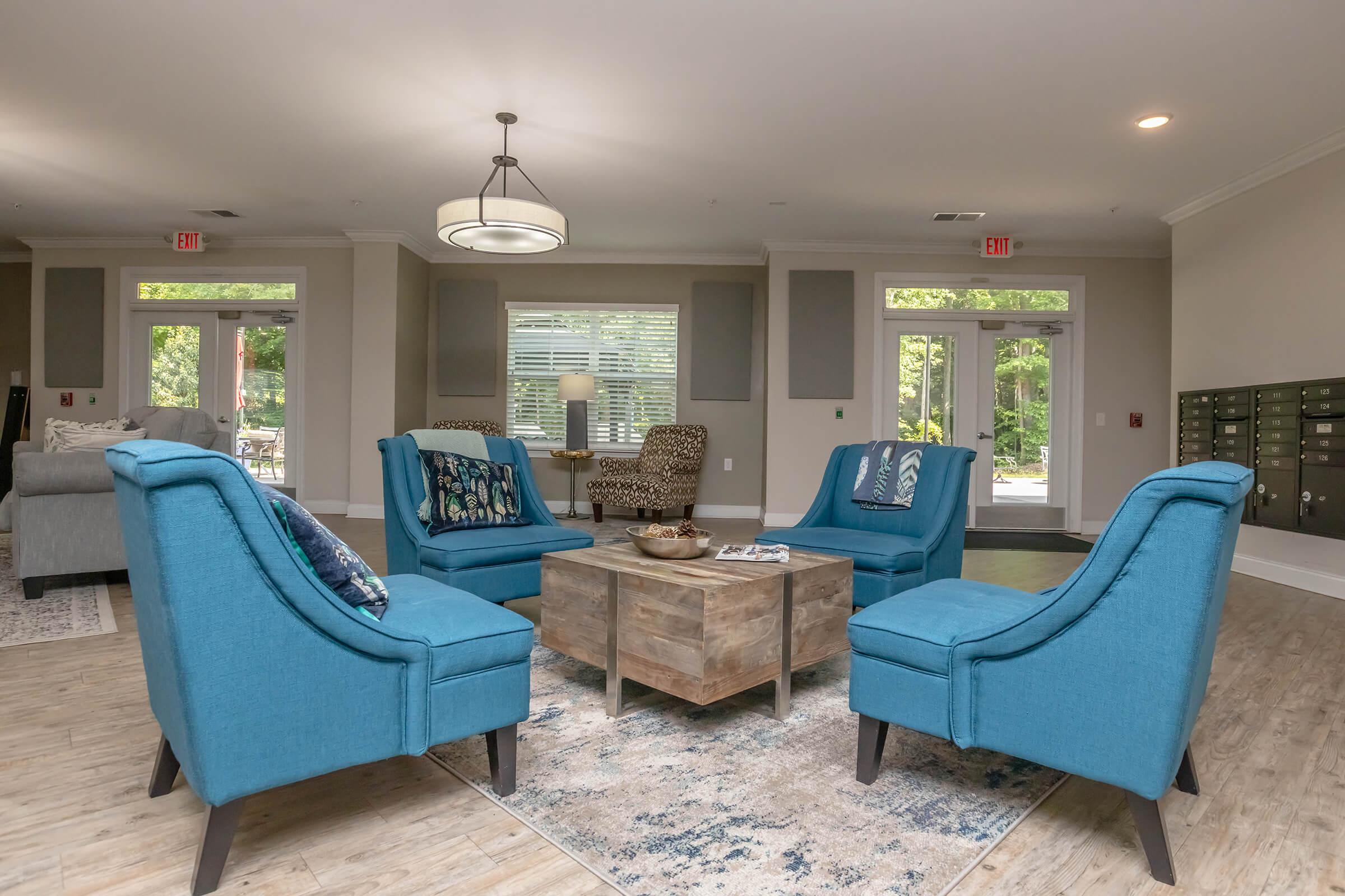 A modern lounge area featuring four stylish blue chairs arranged around a wooden coffee table. The space has large windows that let in natural light, a neutral color palette, and decorative accents, creating a cozy and inviting atmosphere.