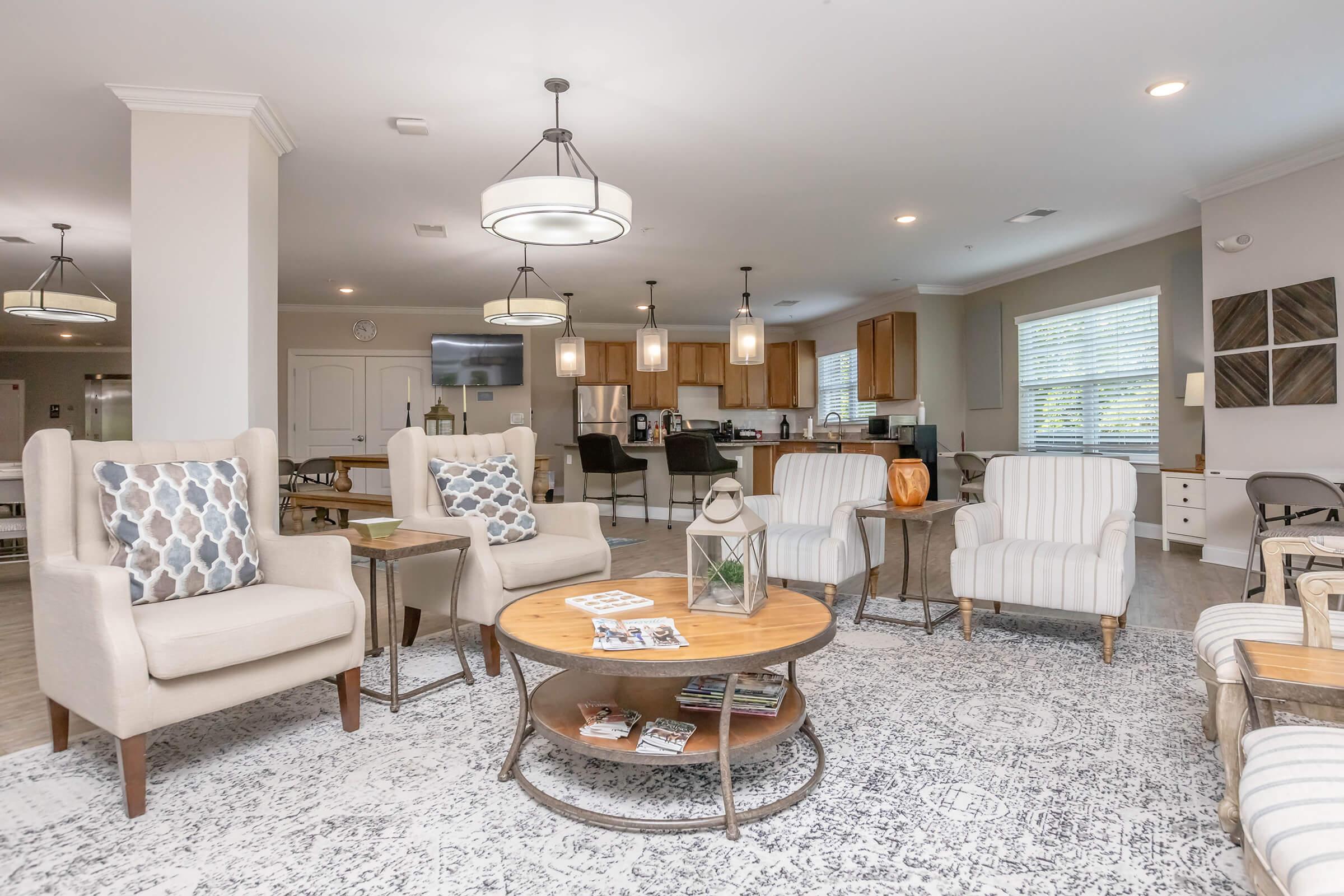 Spacious interior living area featuring light-colored armchairs with patterned cushions, a round wooden coffee table, and a decorative lantern. Soft lighting from pendant lamps and large windows create a warm atmosphere. The background shows a modern kitchen with wooden cabinets.