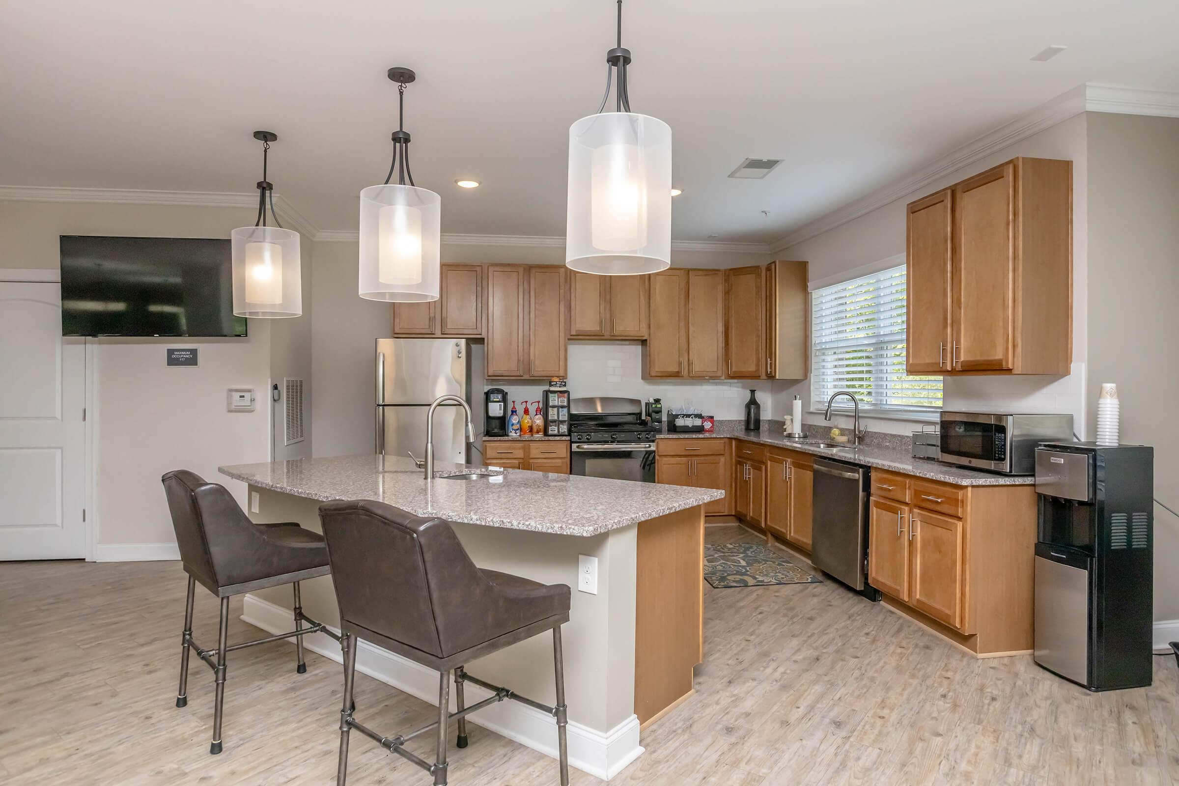 Modern kitchen featuring wooden cabinets, granite countertops, and stainless steel appliances. A spacious island with bar stools provides additional seating. Large windows allow natural light, and stylish pendant lights hang from the ceiling. Appliances include a refrigerator, microwave, and coffee machine.