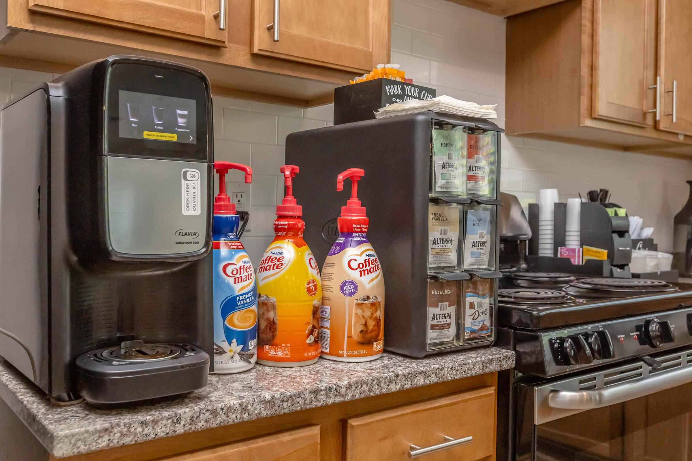 A kitchen countertop featuring a coffee machine, multiple flavored creamers, and a container with coffee supplies. The stove is visible in the background. The appliances and creamers are neatly organized, suggesting a space designed for coffee preparation.