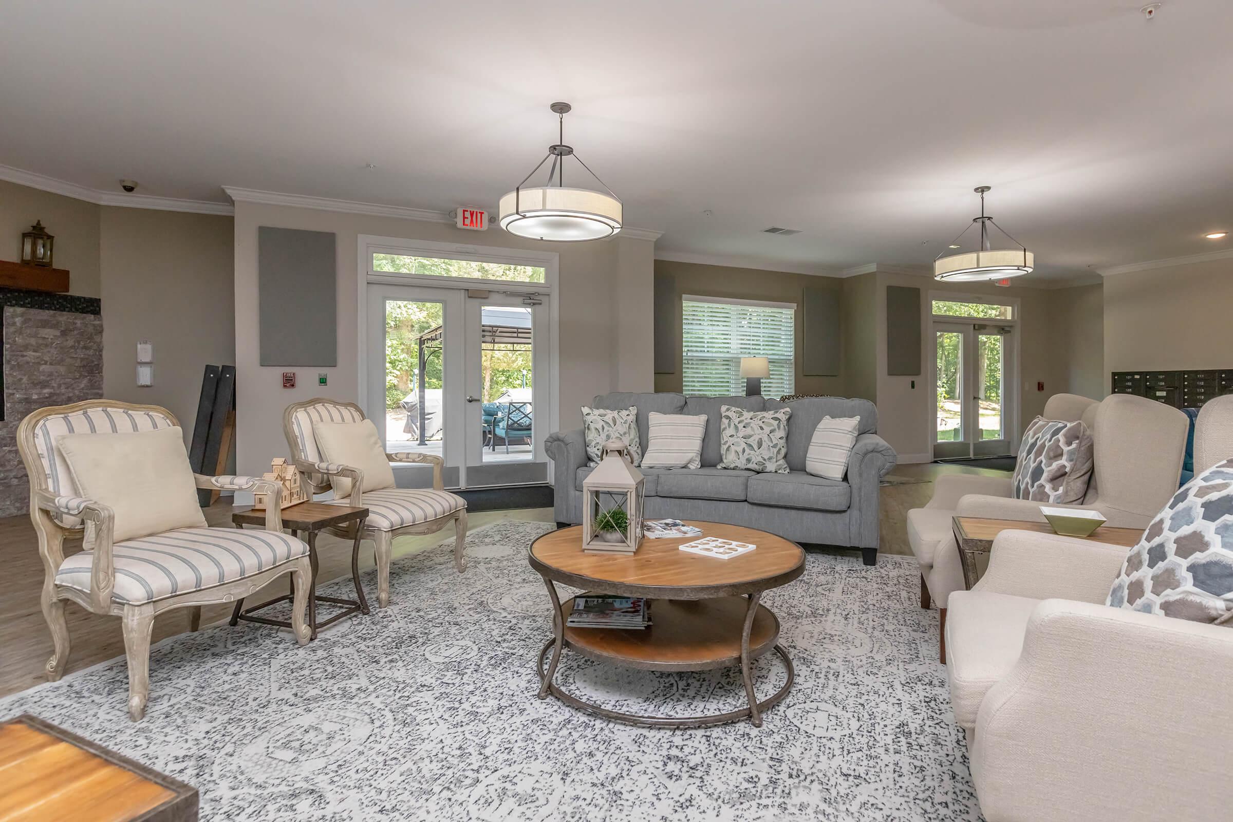 A cozy lounge area featuring a gray sofa with patterned cushions, elegant armchairs, and a round coffee table. Natural light floods in through large windows, highlighting the neutral color palette and decorative accents. The space has a welcoming atmosphere, perfect for relaxation or socialization.