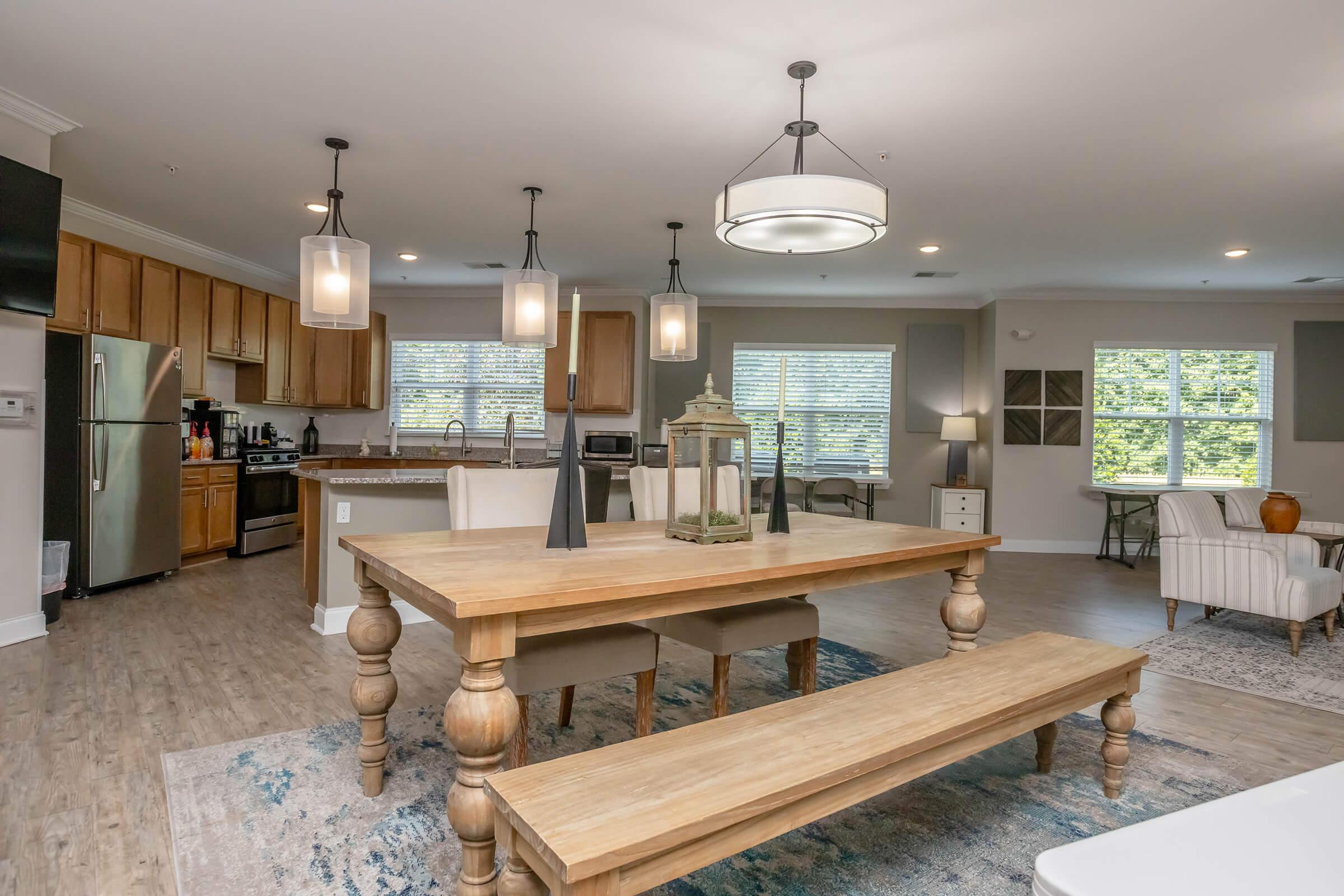 Modern dining area featuring a spacious wooden table with benches, surrounded by stylish chairs. Illuminated by pendant lighting, the open space connects to a contemporary kitchen with stainless steel appliances and ample natural light from large windows. Decor includes a decorative lantern and an area rug.