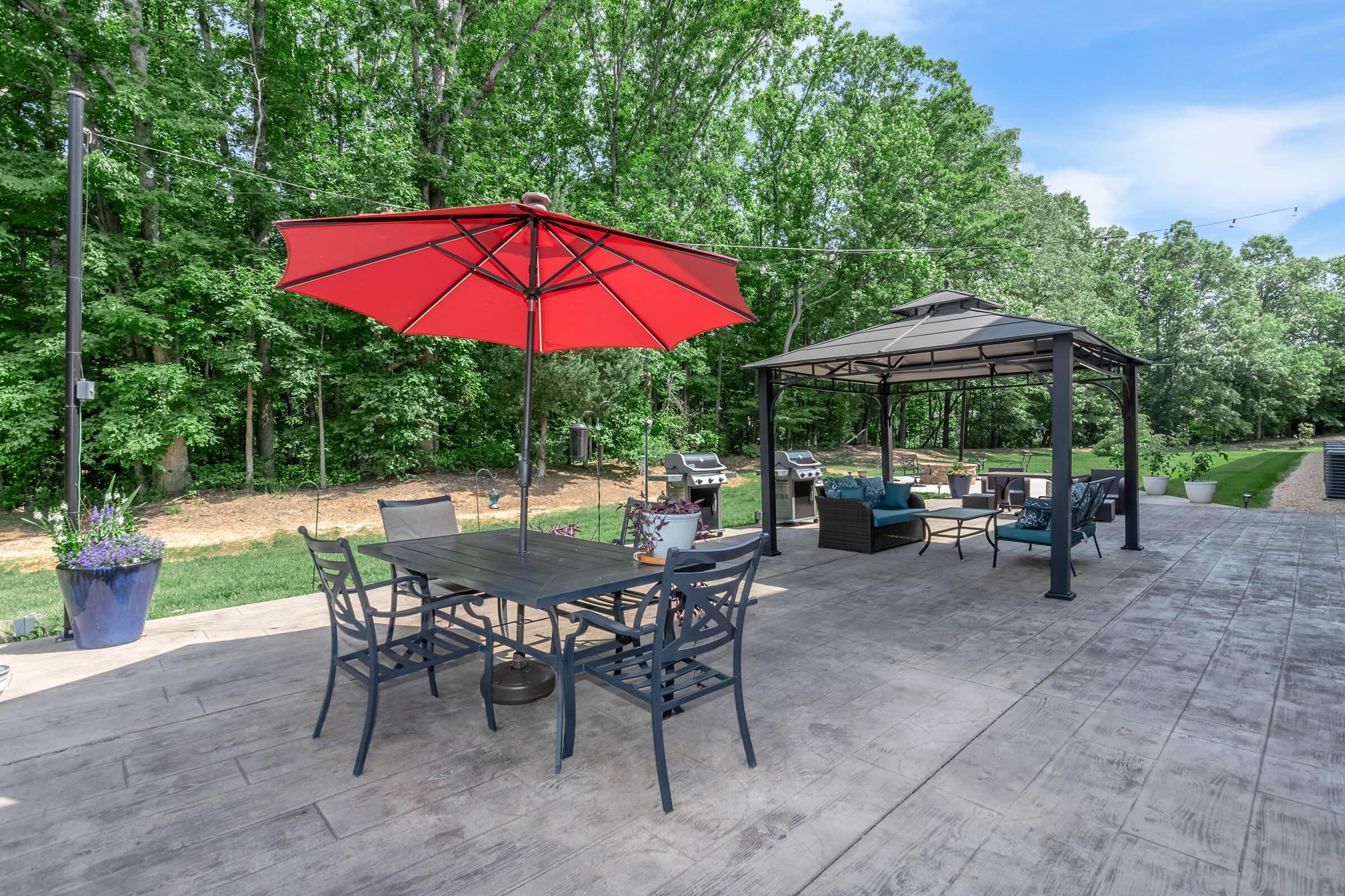 A spacious outdoor patio featuring a large red umbrella over a dining table with chairs, a shaded gazebo with seating, and two grills. Lush greenery surrounds the area, complemented by potted plants and a clear blue sky above. Perfect for relaxation and outdoor gatherings.