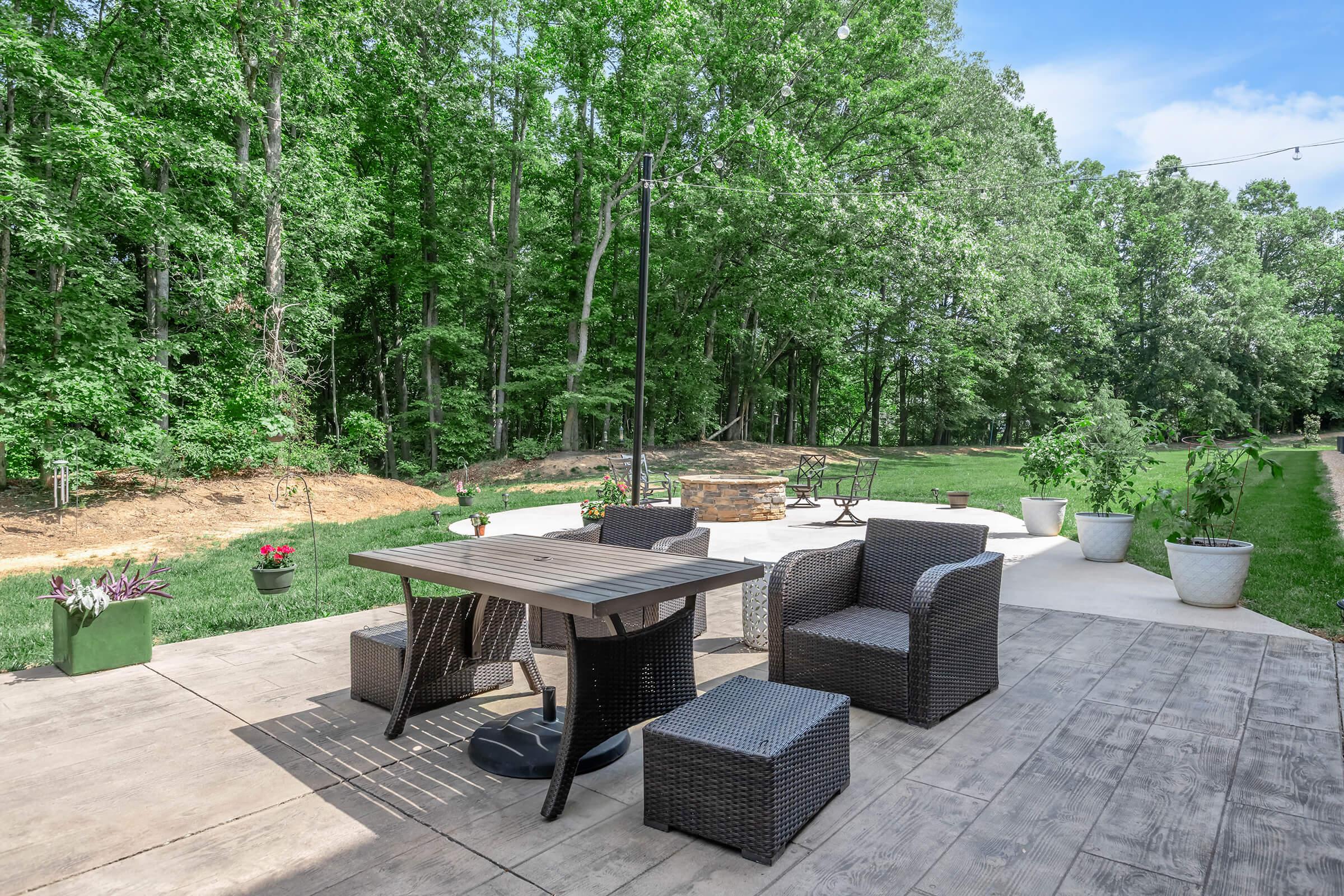 A patio area featuring a wooden table and wicker chairs, surrounded by potted plants. In the background, there are trees and green grass, creating a lush, natural setting. A stone fire pit is visible nearby, along with a pathway leading further into the garden.