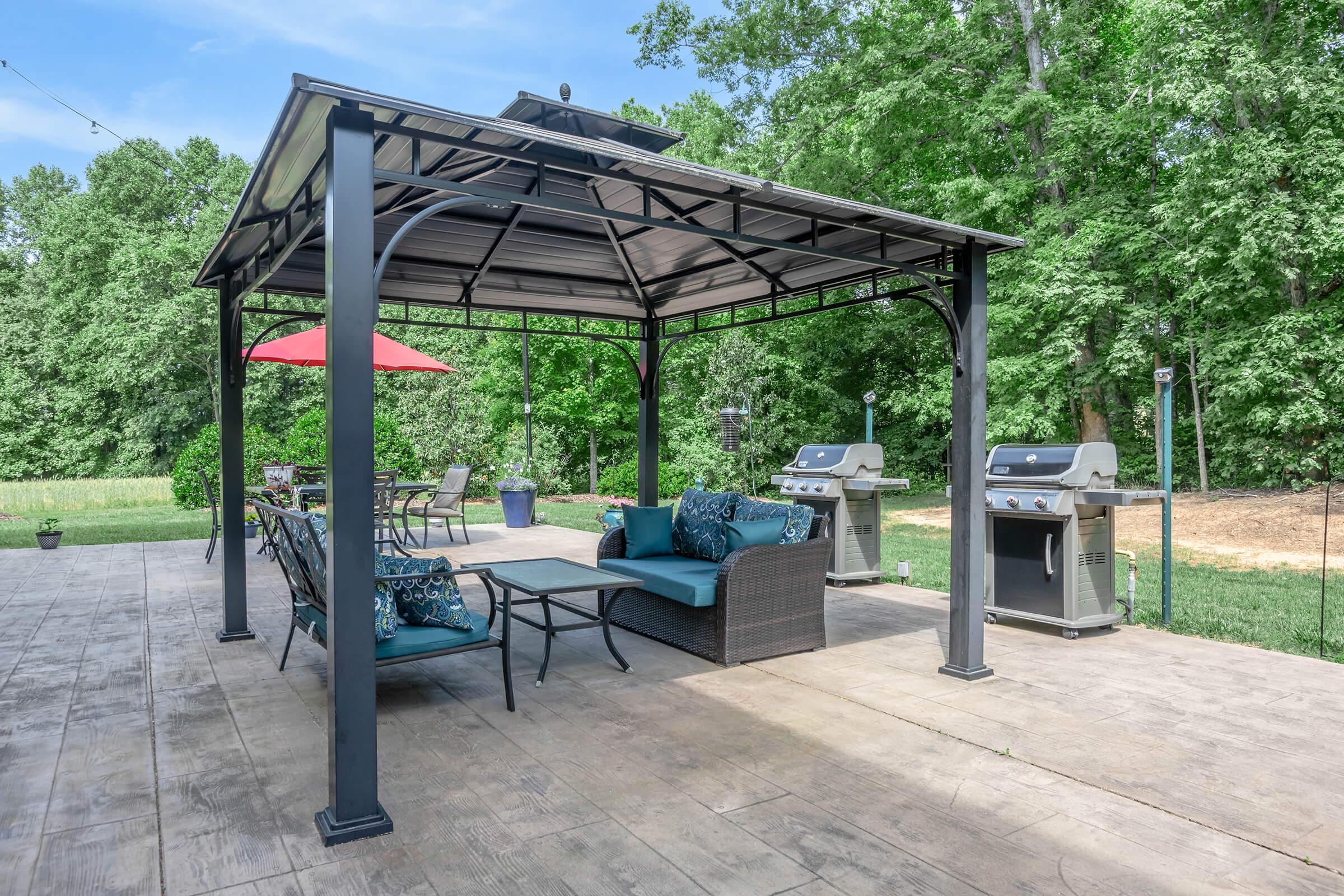 A spacious outdoor patio with a gazebo featuring seating and cushions, surrounded by greenery. There are two grilling stations on either side, and a large table with chairs. A red umbrella provides shade, enhancing the inviting atmosphere for outdoor gatherings.