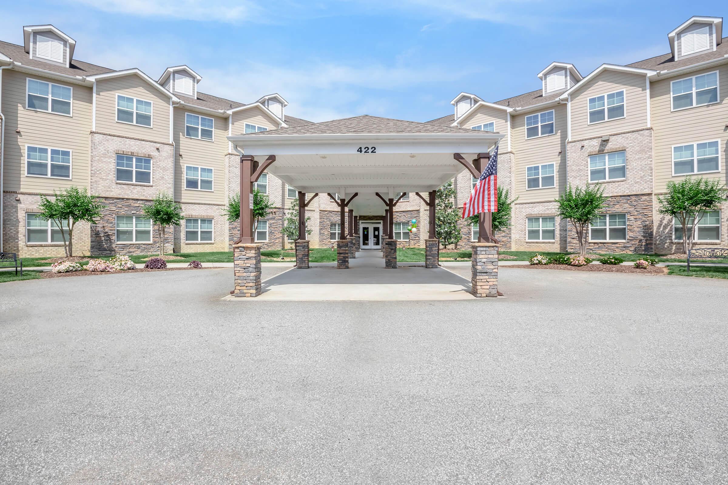 A welcoming entrance of a modern residential building, featuring a covered walkway supported by stone columns. The facade includes multiple windows and a neat landscaping with shrubs. An American flag is displayed, adding a patriotic element to the scene.