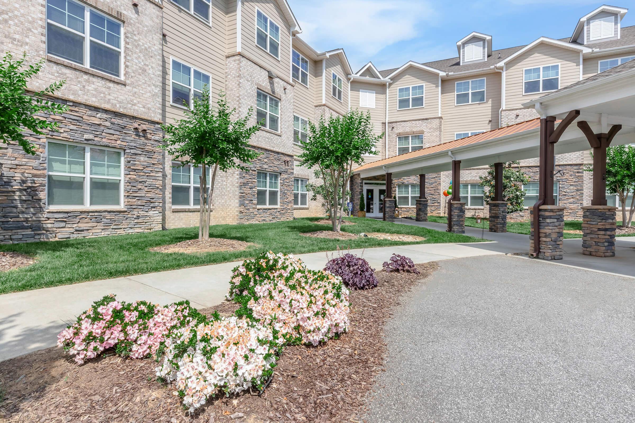 A landscaped area featuring colorful flower beds with pink and purple blooms in front of a large multi-story residential building. The building has a mix of brick and siding, large windows, and a covered entrance with trees and greenery surrounding it.