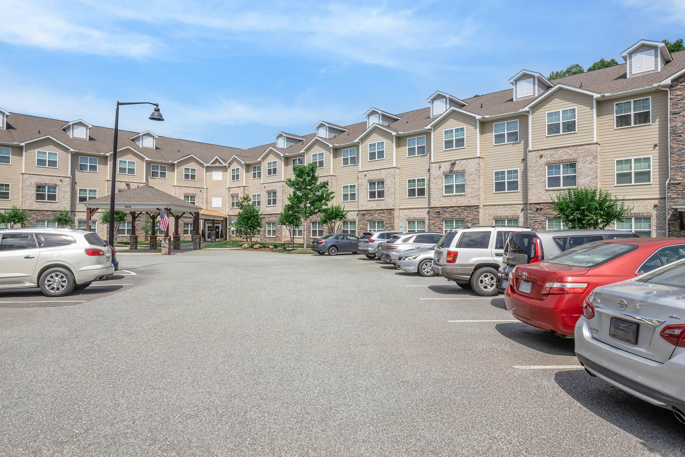 A view of an apartment complex featuring multiple two-story buildings with stone accents, surrounded by a parking lot with several cars. There are trees and greenery in the area, with clear blue skies overhead, creating a welcoming residential environment.