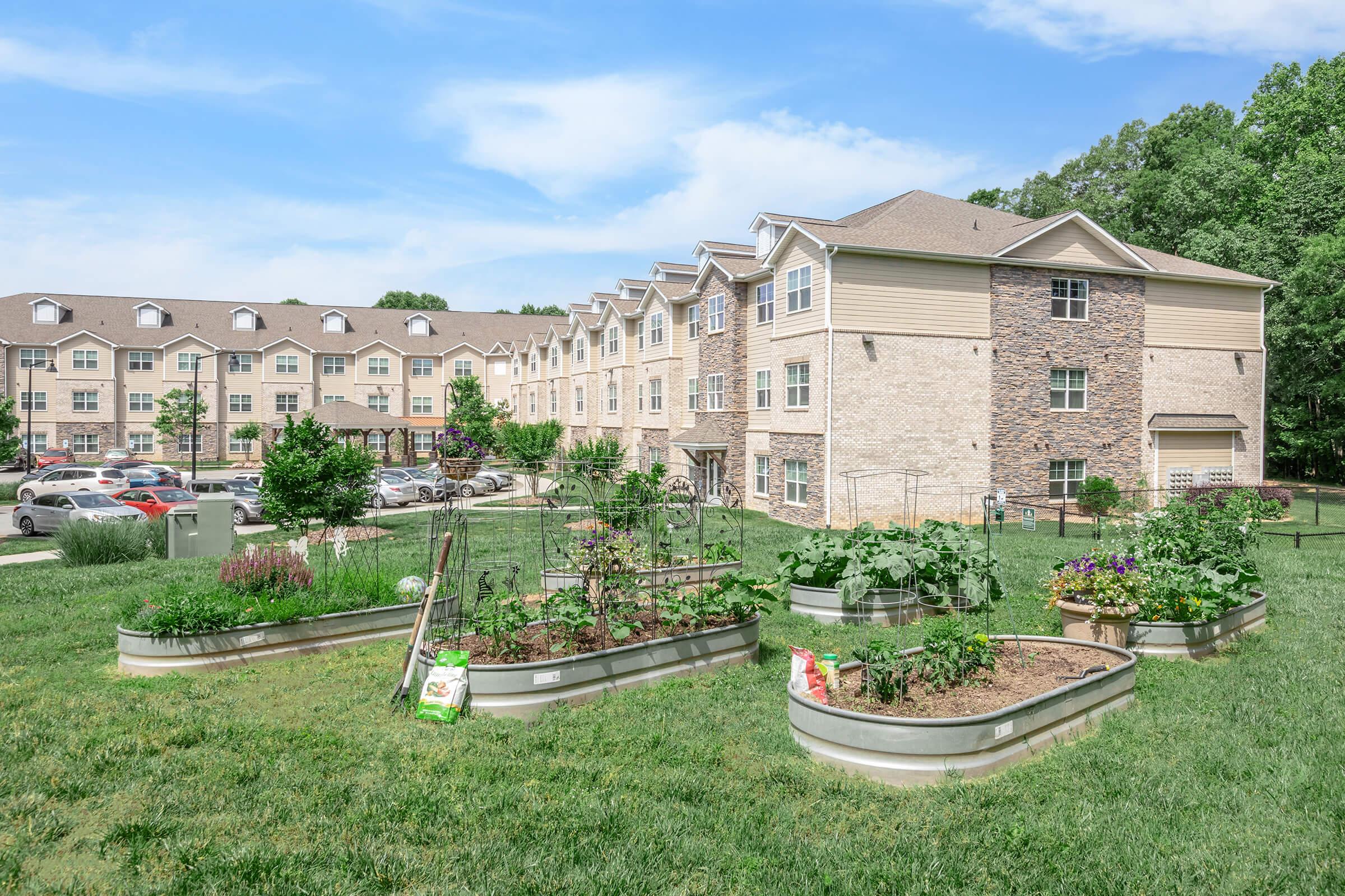 A landscaped area featuring raised garden beds filled with various plants in the foreground, with a multi-story apartment building in the background. The scene is set in a grassy area under a blue sky, showcasing a mix of greenery and an organized apartment complex.