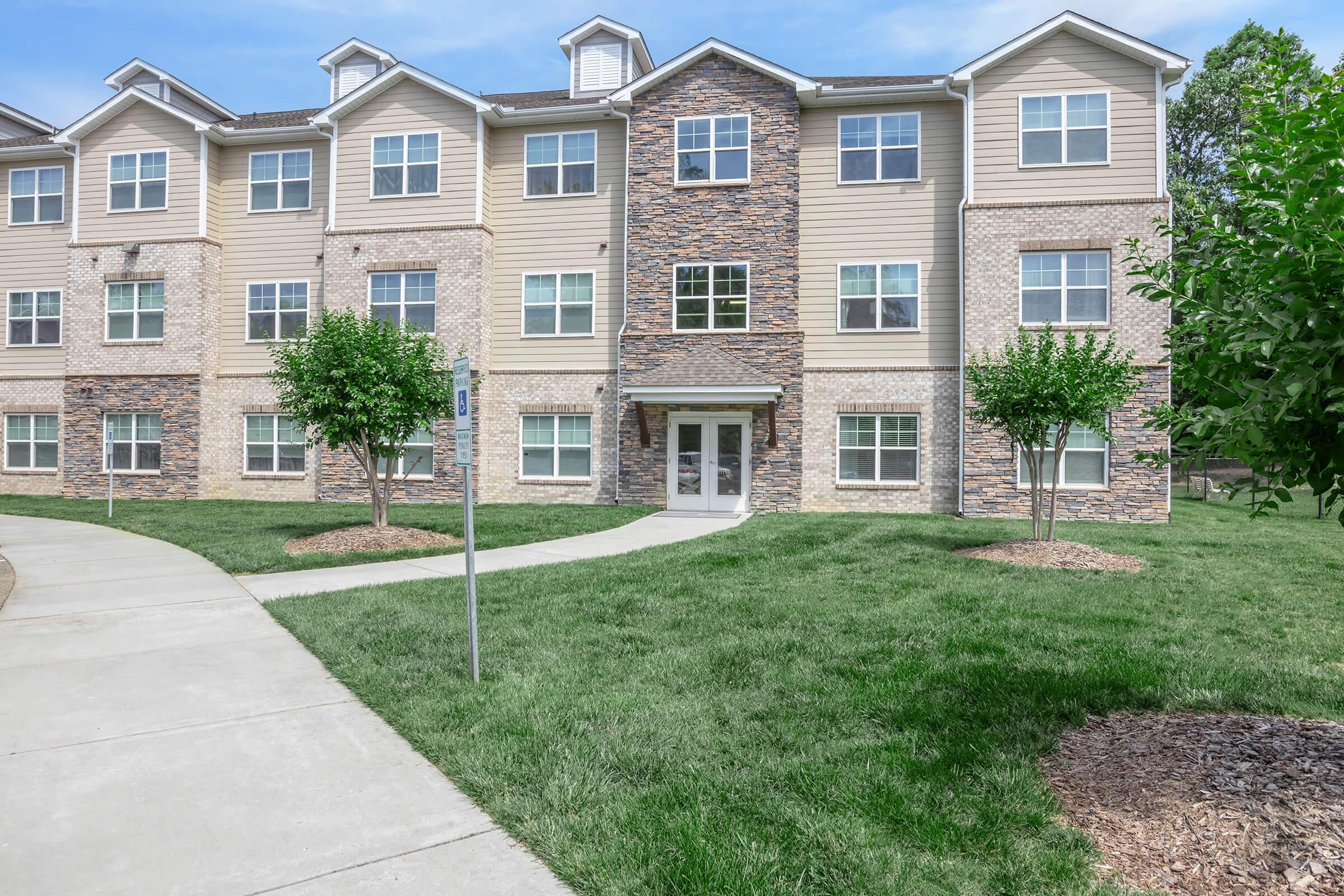 A three-story residential building featuring a combination of stone and siding exterior. It has multiple windows, a front entrance with a small porch, and greenery around, including lawn and small trees. A walking path leads towards the entrance, providing access to the building.