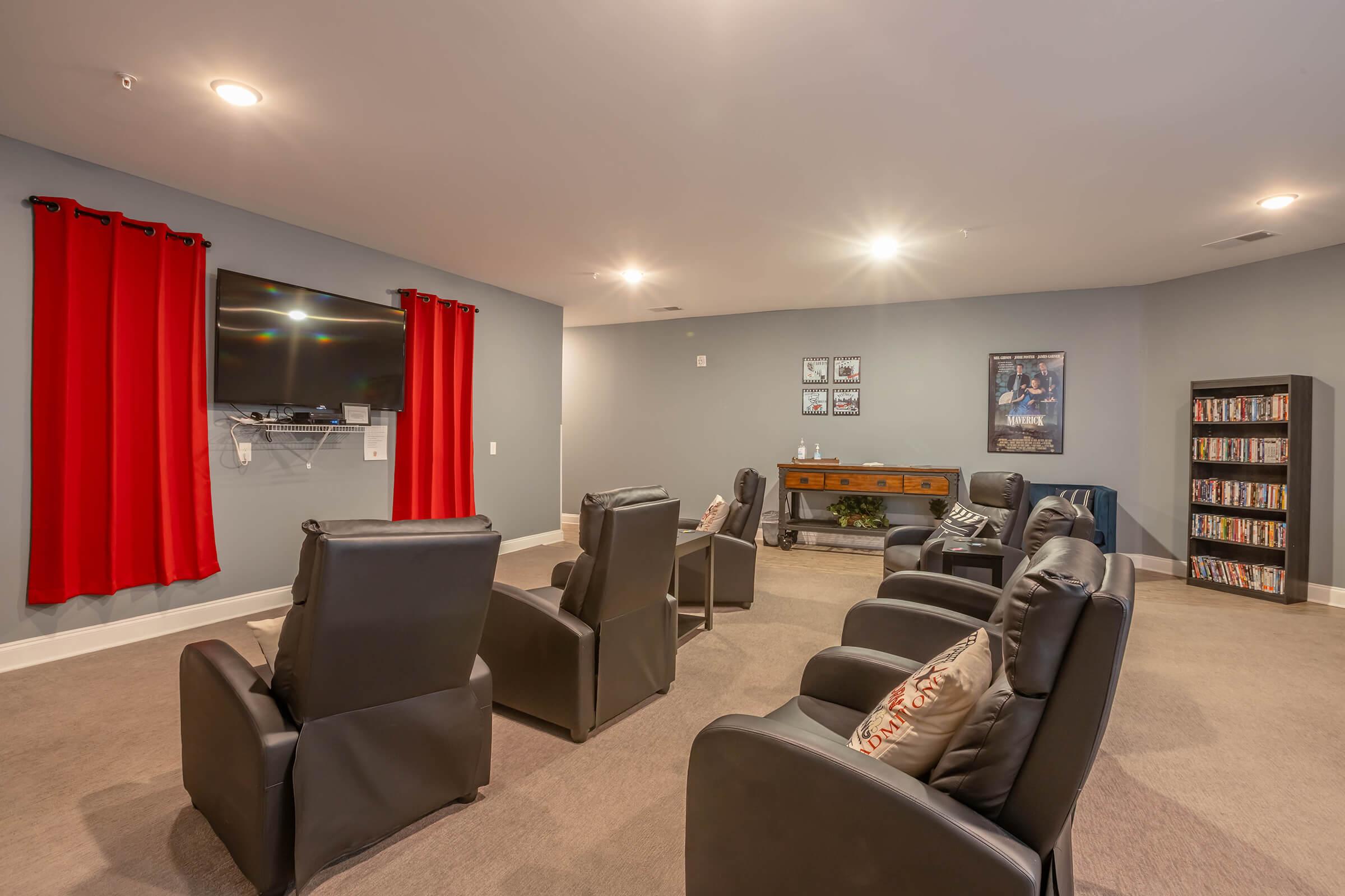 A cozy entertainment room featuring several black reclining chairs arranged for viewing a wall-mounted television. The room has light gray walls, dim lighting, and red curtains. There's a wooden console table against one wall, adorned with picture frames, and a shelf filled with DVDs, creating a comfortable movie-watching atmosphere.