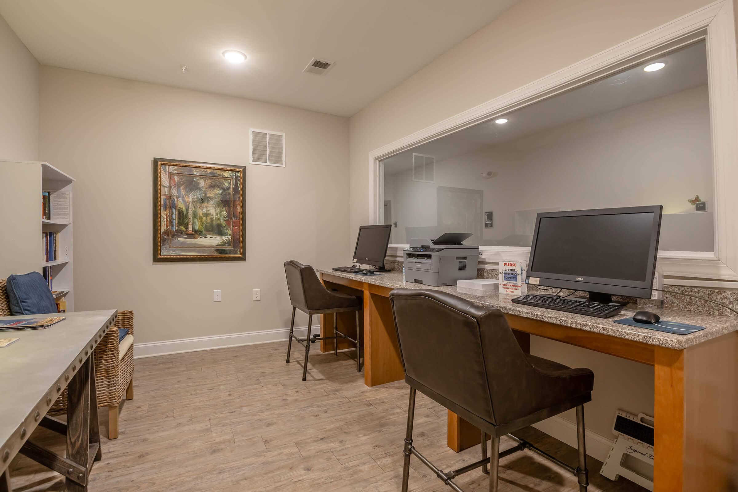 A modern office space featuring two desks with computers and a printer. One wall has a framed painting, while another wall includes a bookshelf. The flooring is wooden, and the walls are painted in neutral tones, creating a professional and inviting atmosphere.