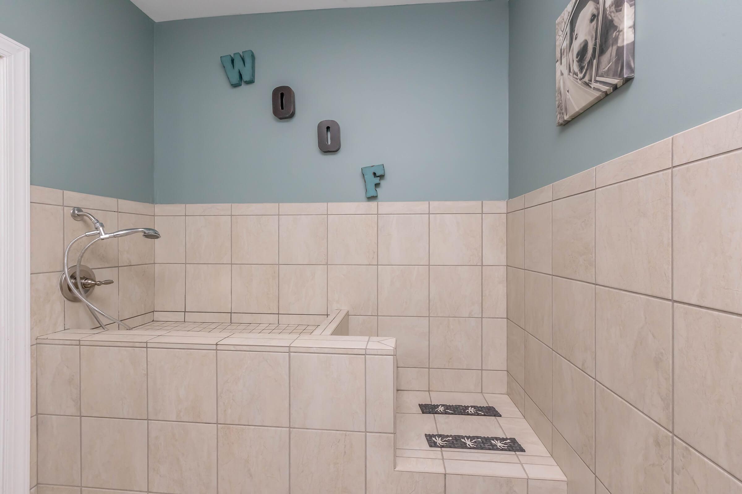 A modern bathroom featuring a tiled shower with a built-in bench. The walls are painted light blue, and decorative letters spelling "WOOF" are mounted on the wall. There are patterned mats in front of the shower area, and a decorative wall art piece is visible. The overall design is clean and contemporary.