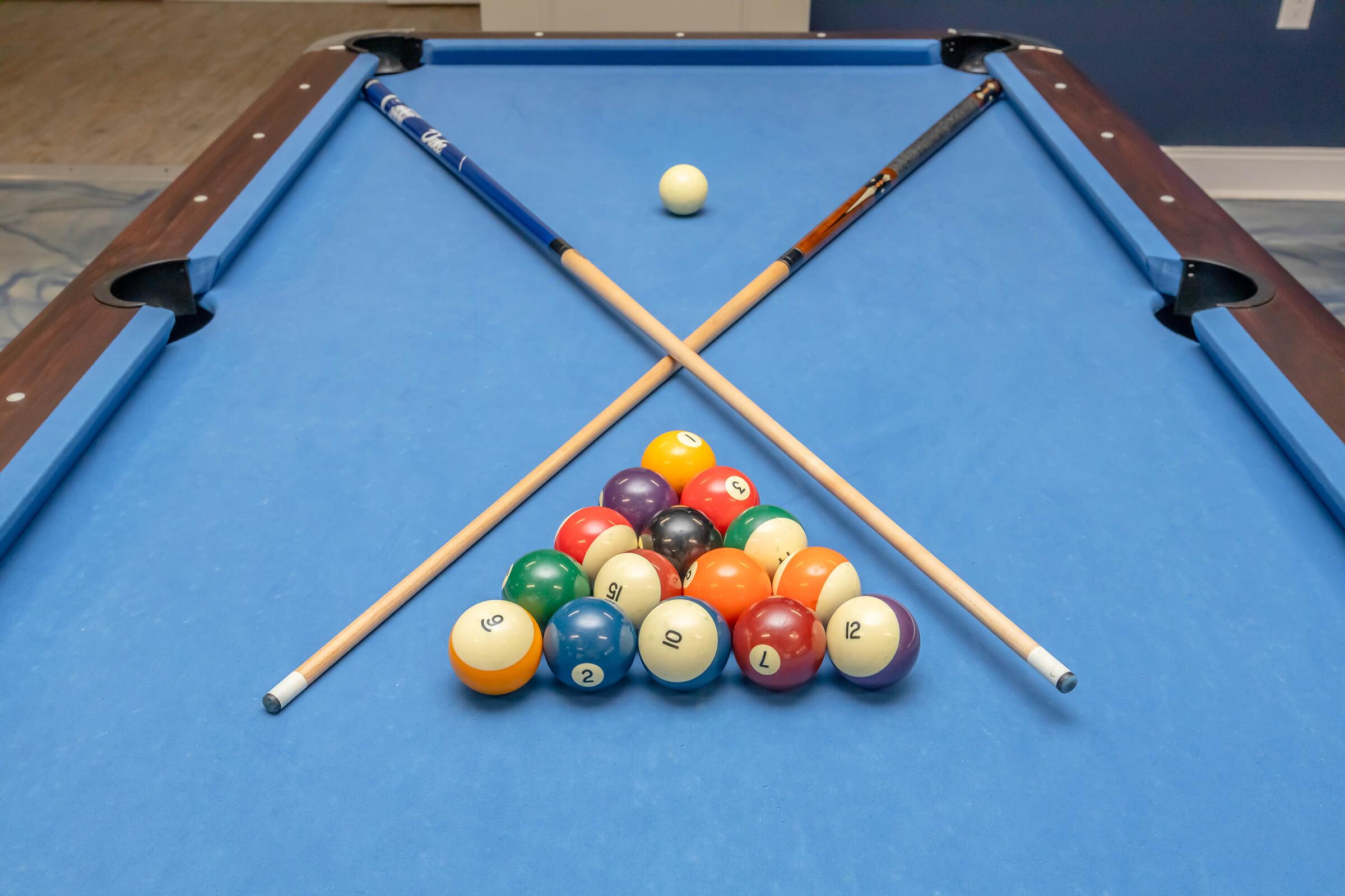 A pool table with a blue felt surface featuring a triangle rack of colored billiard balls arranged in a group. Two pool cues are placed diagonally across the table, and a cue ball is positioned nearby, ready for play.
