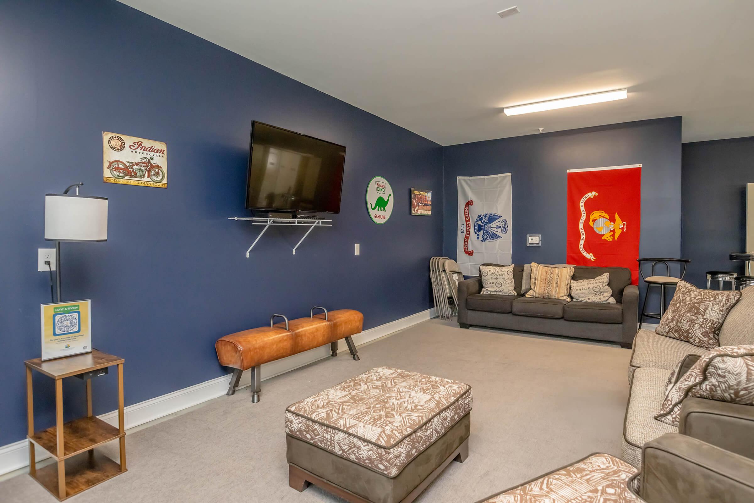 A cozy living room featuring a dark blue wall, a flat-screen TV mounted on a shelf, and various decorative items including flags and vintage signs. Comfortable sofas and chairs are arranged around an ottoman, with a lamp and side table providing additional lighting and space.