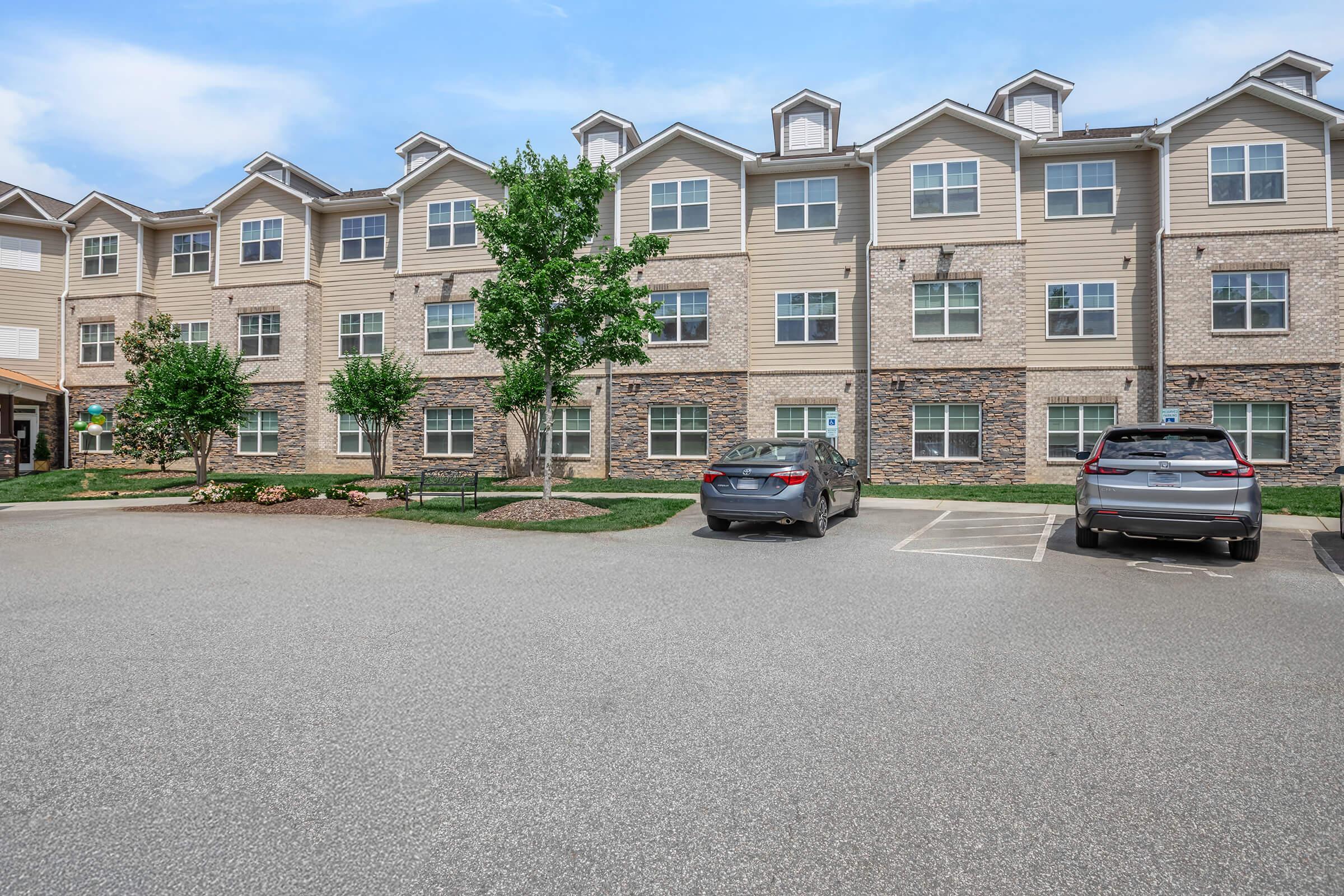 A multi-story apartment building with a brick and siding exterior. The front features several windows and a small parking area with two cars. There are trees and landscaped areas surrounding the building, and the sky is clear and blue.
