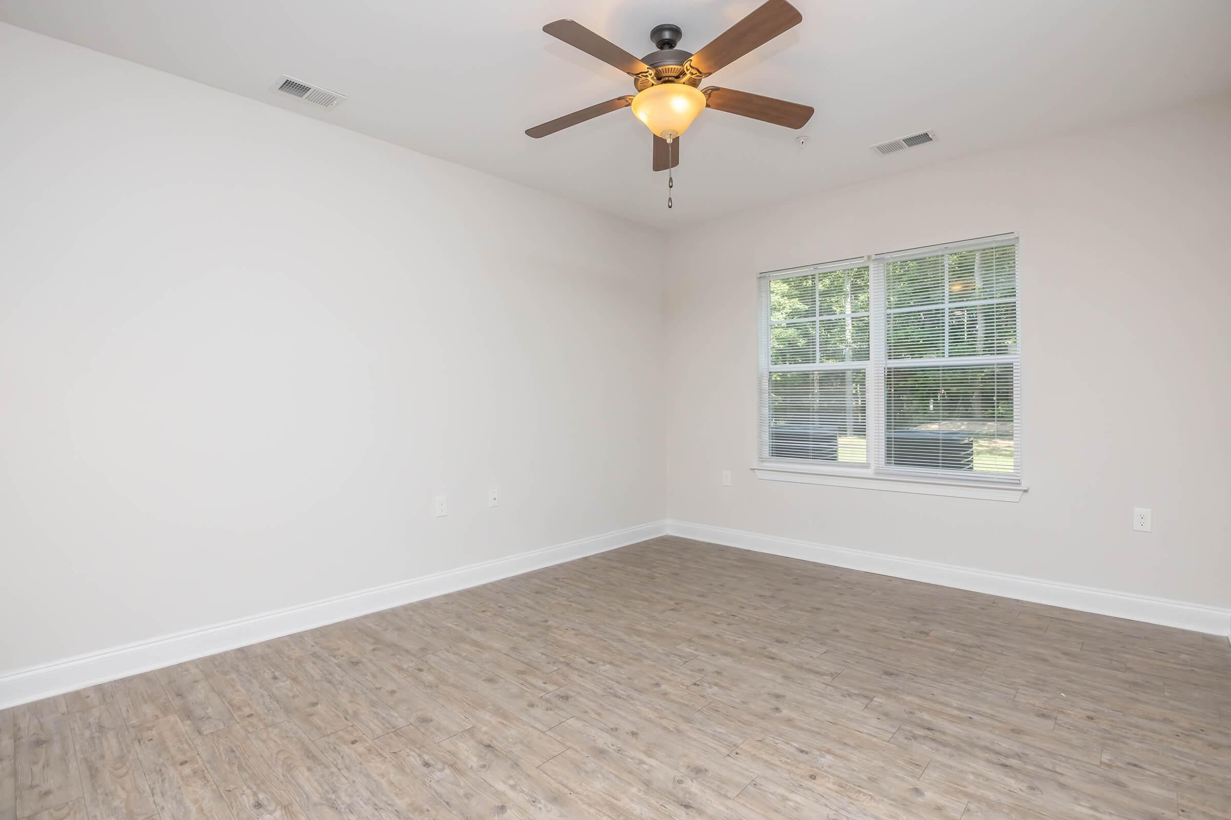 A spacious, empty room with light-colored walls and a wooden floor. A ceiling fan with a light fixture is centered above, and a window with multiple panes lets in natural light. The overall atmosphere is bright and inviting, making it suitable for various uses.