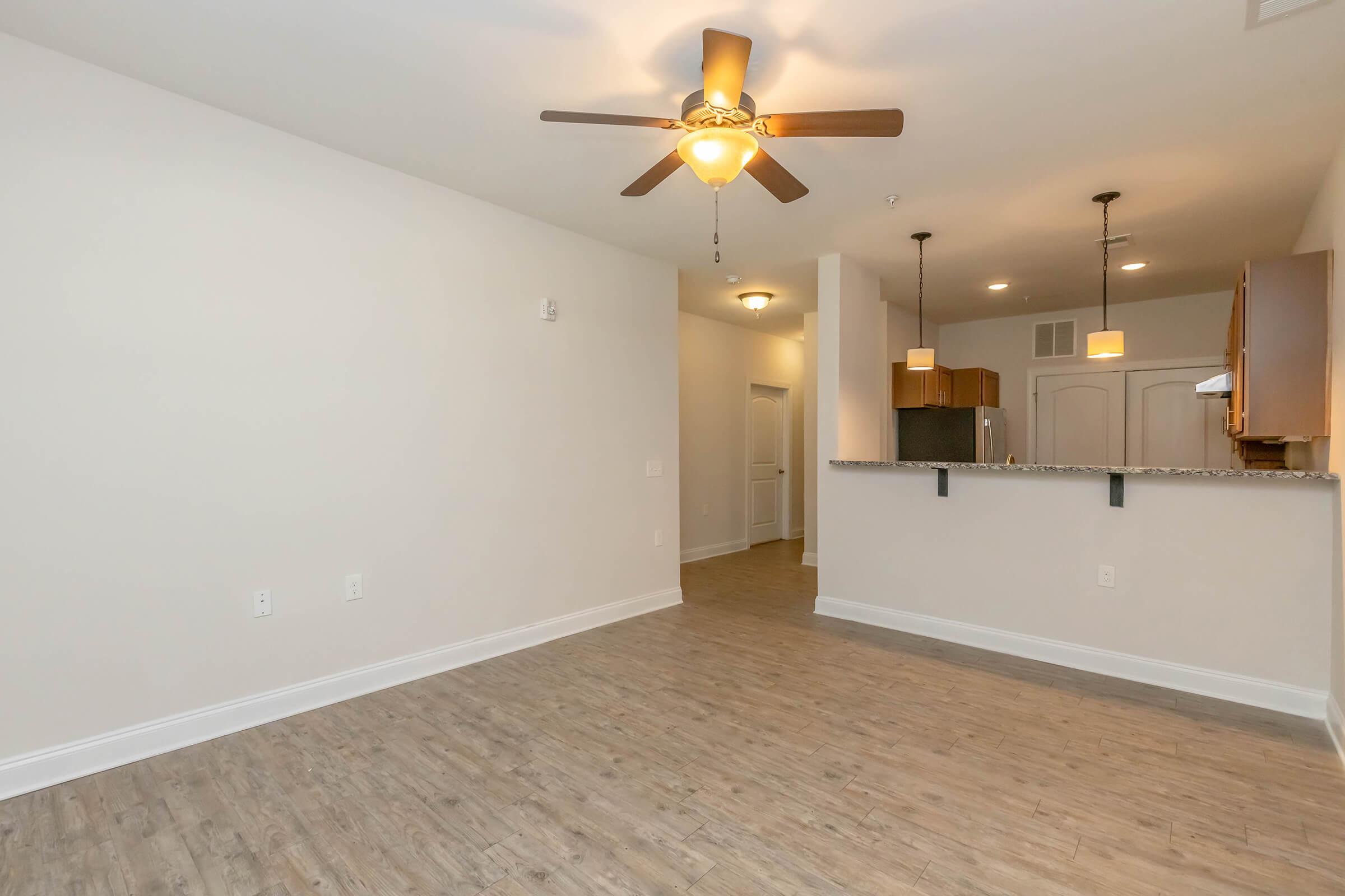 A spacious, well-lit living area featuring a ceiling fan, light hardwood flooring, and a neutral color scheme. The room includes a bar counter leading to a kitchen area with dark wooden cabinets, creating an inviting atmosphere. The layout is open, enhancing the feeling of space and comfort.