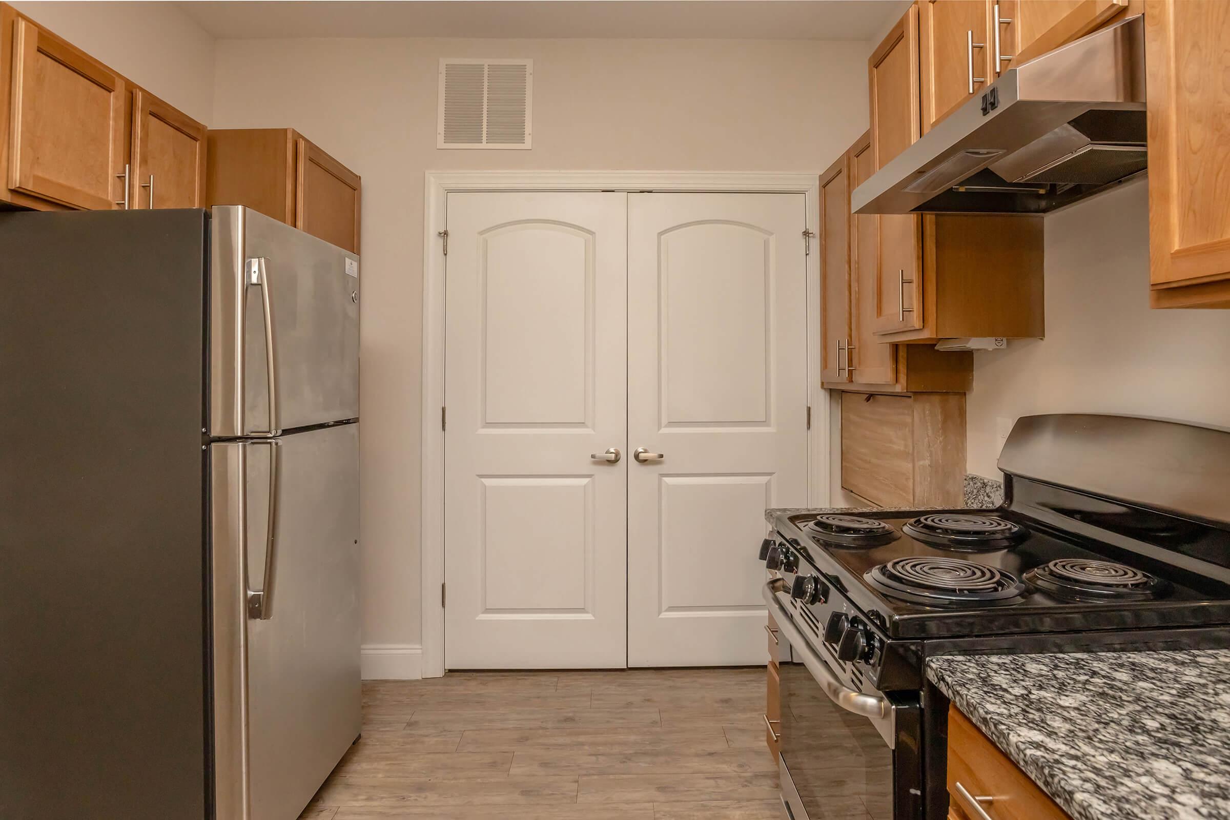 A modern kitchen featuring wooden cabinets, a stainless steel refrigerator, and a black stove with four burners. The kitchen has a granite countertop and light-colored walls. Two white double doors are visible at the end of the kitchen, providing access to another room.