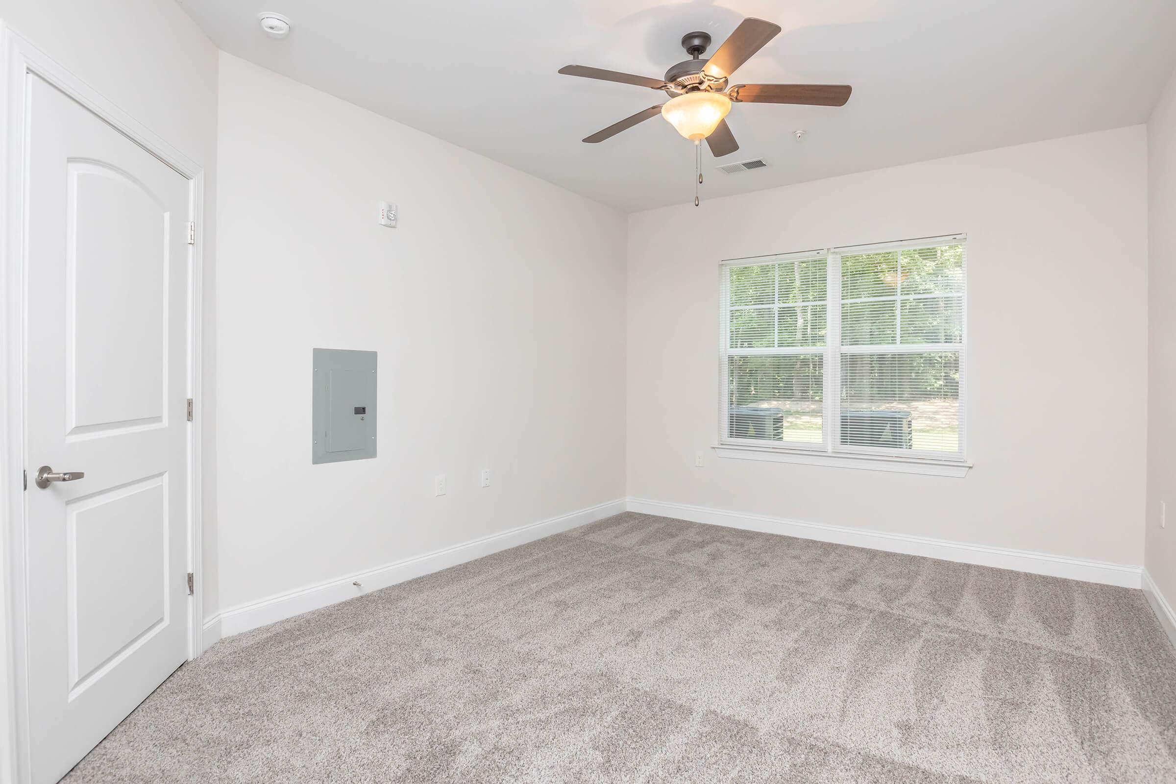 A well-lit, empty room featuring light-colored walls and plush carpet. It includes a ceiling fan, a window with multiple panes allowing natural light, and a wall-mounted electrical panel. The space is clean, with no furniture, offering a blank canvas for decoration or furnishing.