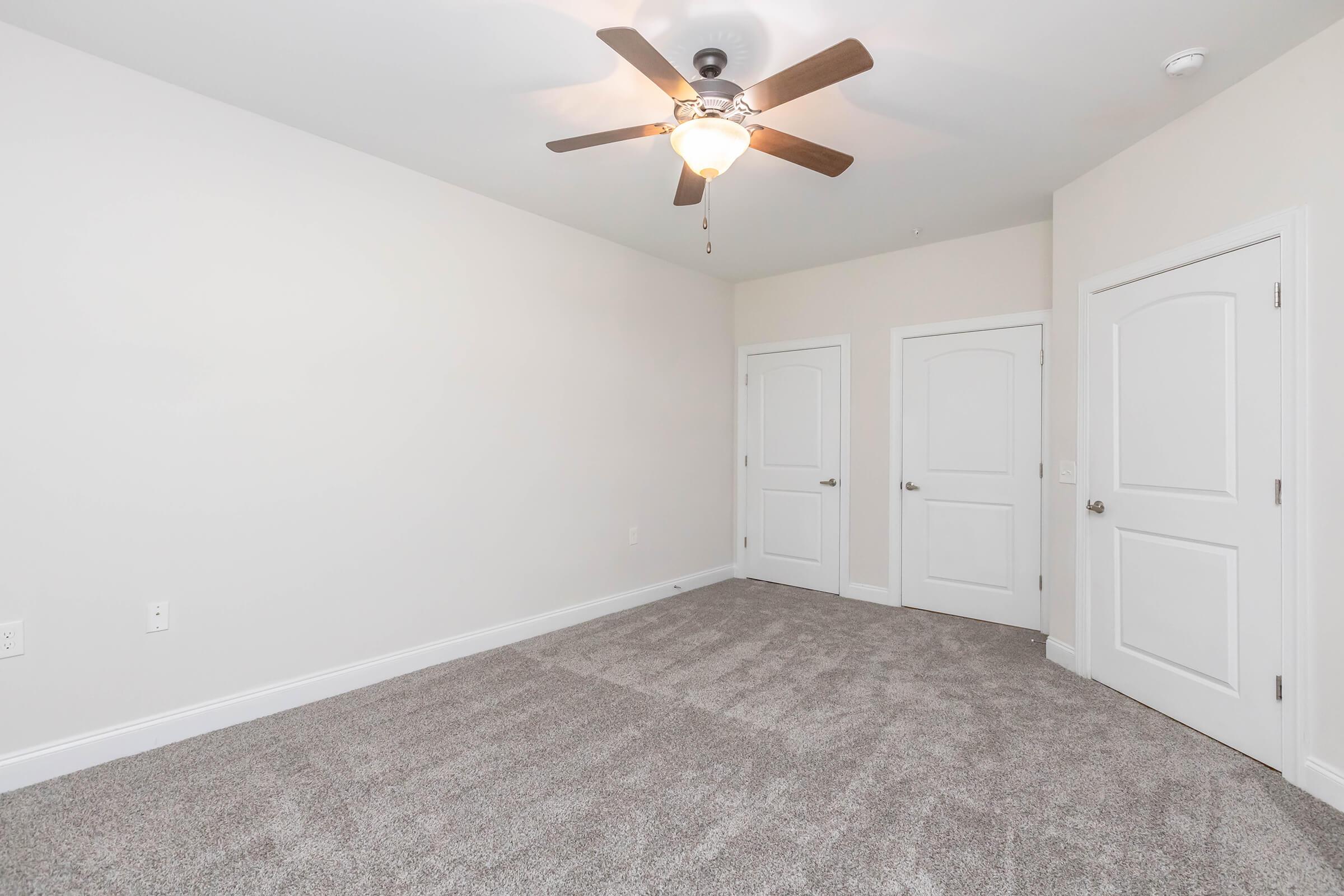 A light-colored room with plush gray carpet, featuring a ceiling fan, two white doors on the right, and a plain white wall. The space is unfurnished, providing a clean and simple atmosphere, suitable for various uses.