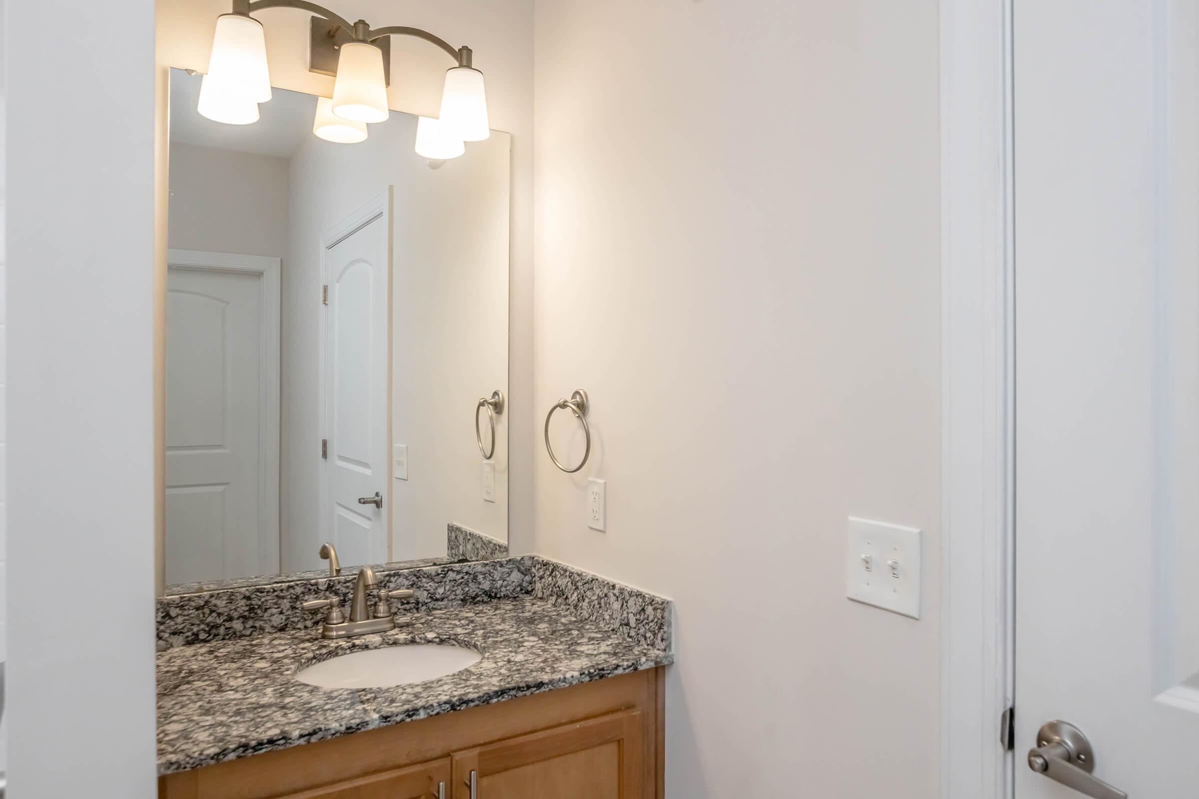 A bathroom vanity with a granite countertop, a round sink, and a wall-mounted mirror illuminated by three hanging light fixtures. The walls are light-colored, and there's a towel ring beside the sink. A closed white door is visible in the background, leading to another room.