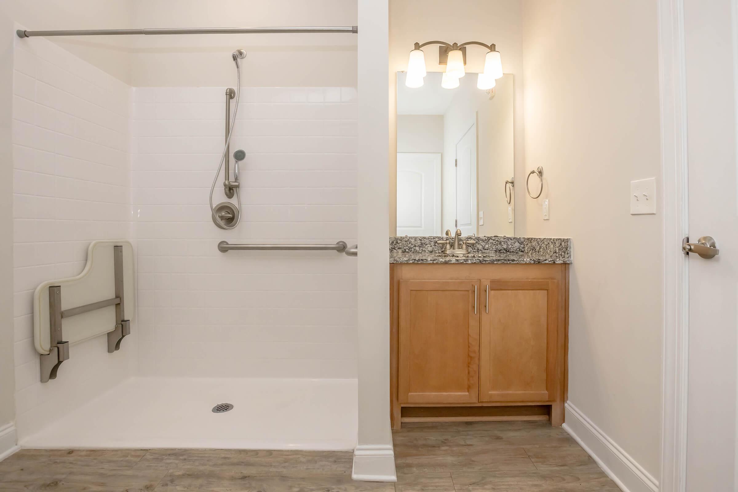 A modern bathroom featuring a walk-in shower with grab bars and a folding shower seat on the left. On the right, there is a wooden vanity with a granite countertop and a mirror above, illuminated by wall-mounted lights. The walls are painted in a light color, creating a spacious feel.