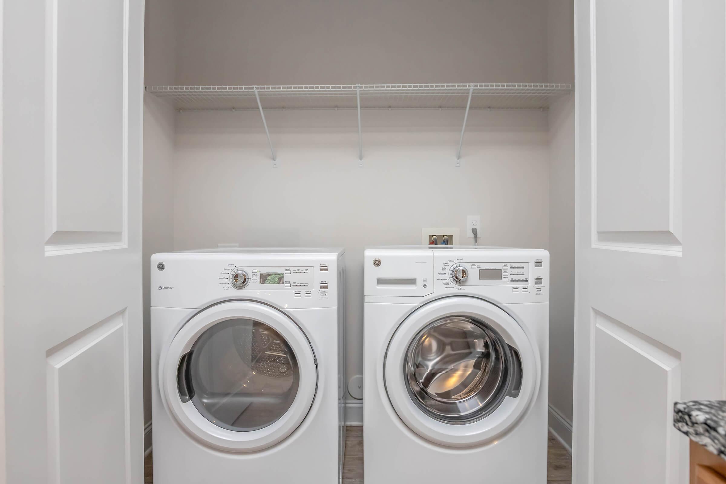 A compact laundry area featuring two white washing machines side by side, with a rack above for storage. The space is neatly organized and bright, with light-colored walls and flooring.