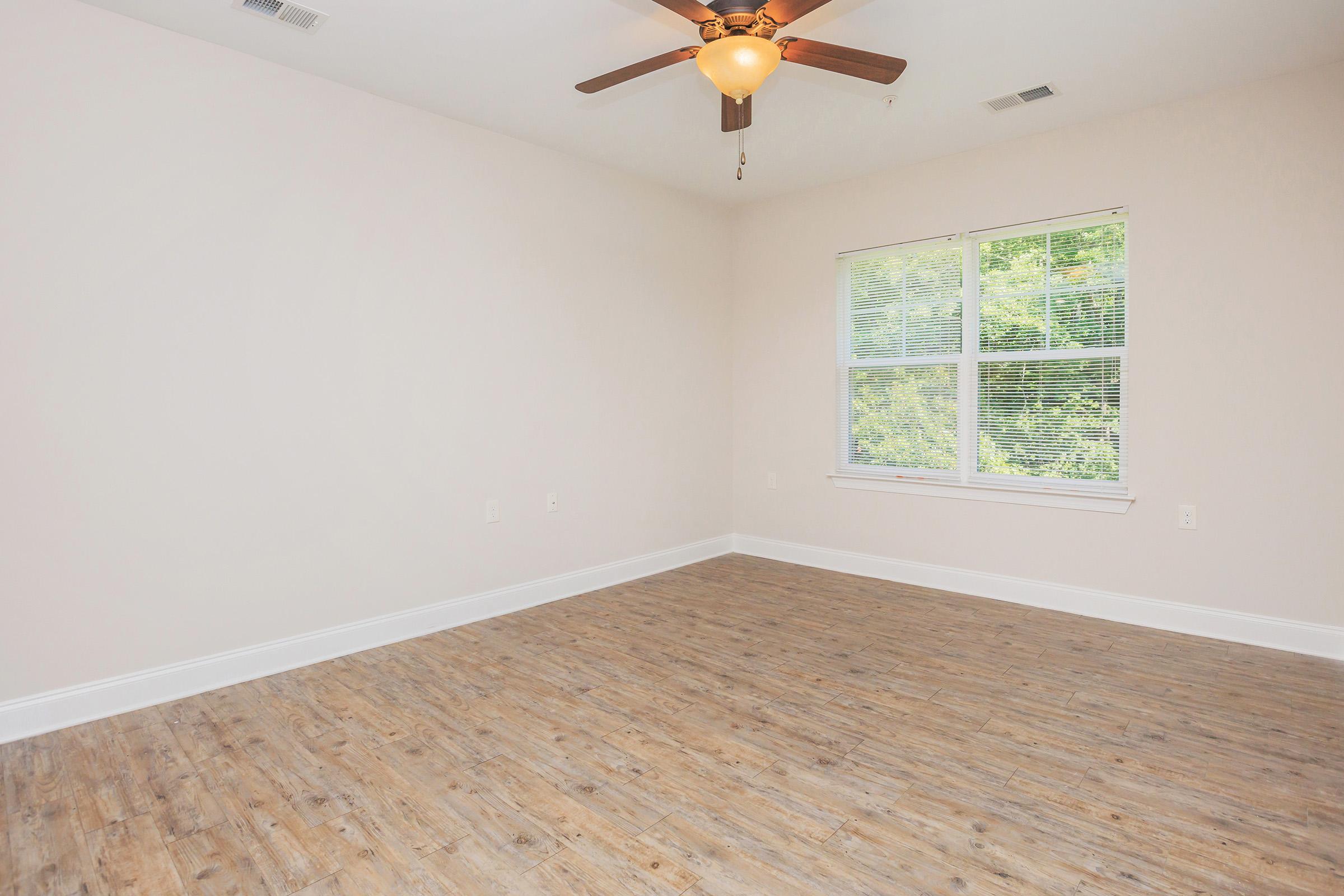 Empty room with light-colored walls and wooden flooring. A ceiling fan is mounted in the center, and there is a large window with multiple panes, allowing natural light to enter and providing a view of greenery outside. The space is bright and spacious with no furniture present.