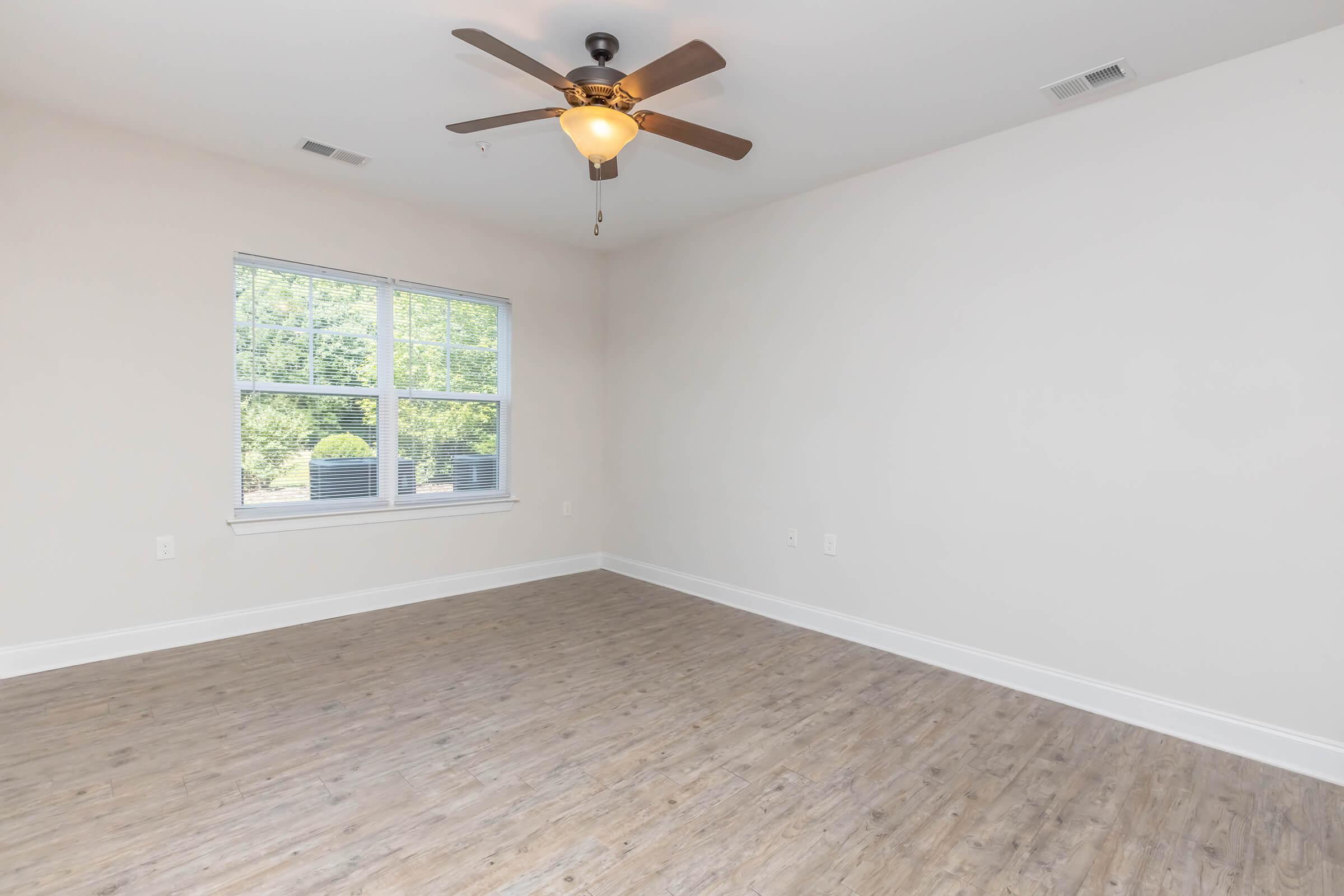 A spacious empty room featuring light wood-like flooring, a ceiling fan with a light fixture, and a large window that lets in natural light. The walls are painted in a soft neutral color, creating an airy and open atmosphere.