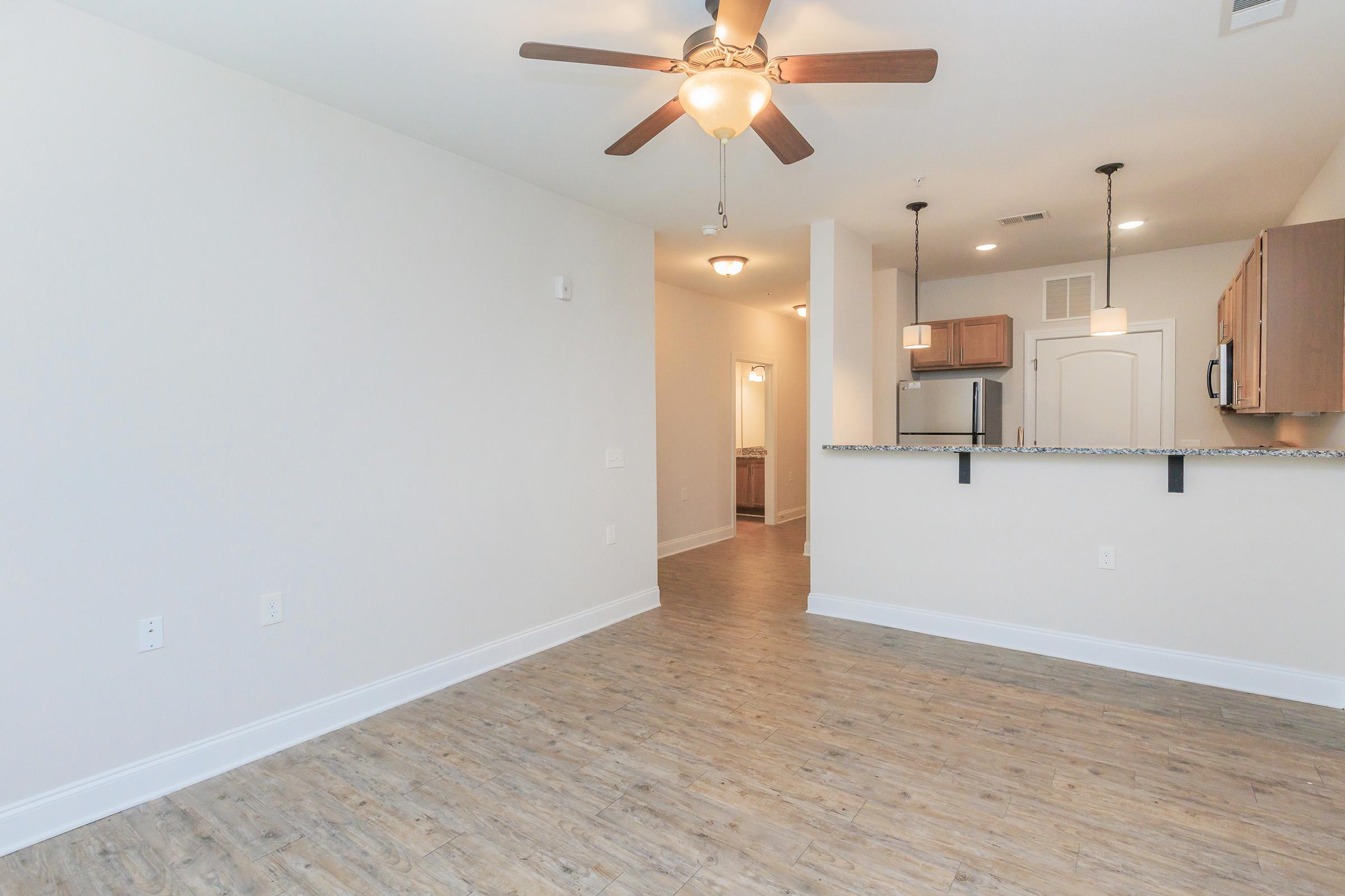 A spacious, modern living area with light wood flooring, a ceiling fan, and neutral-colored walls. There is an open kitchen area with wooden cabinetry and a breakfast bar. A hallway leads to a bathroom, visible through an open door. Natural light fills the room, creating a welcoming atmosphere.